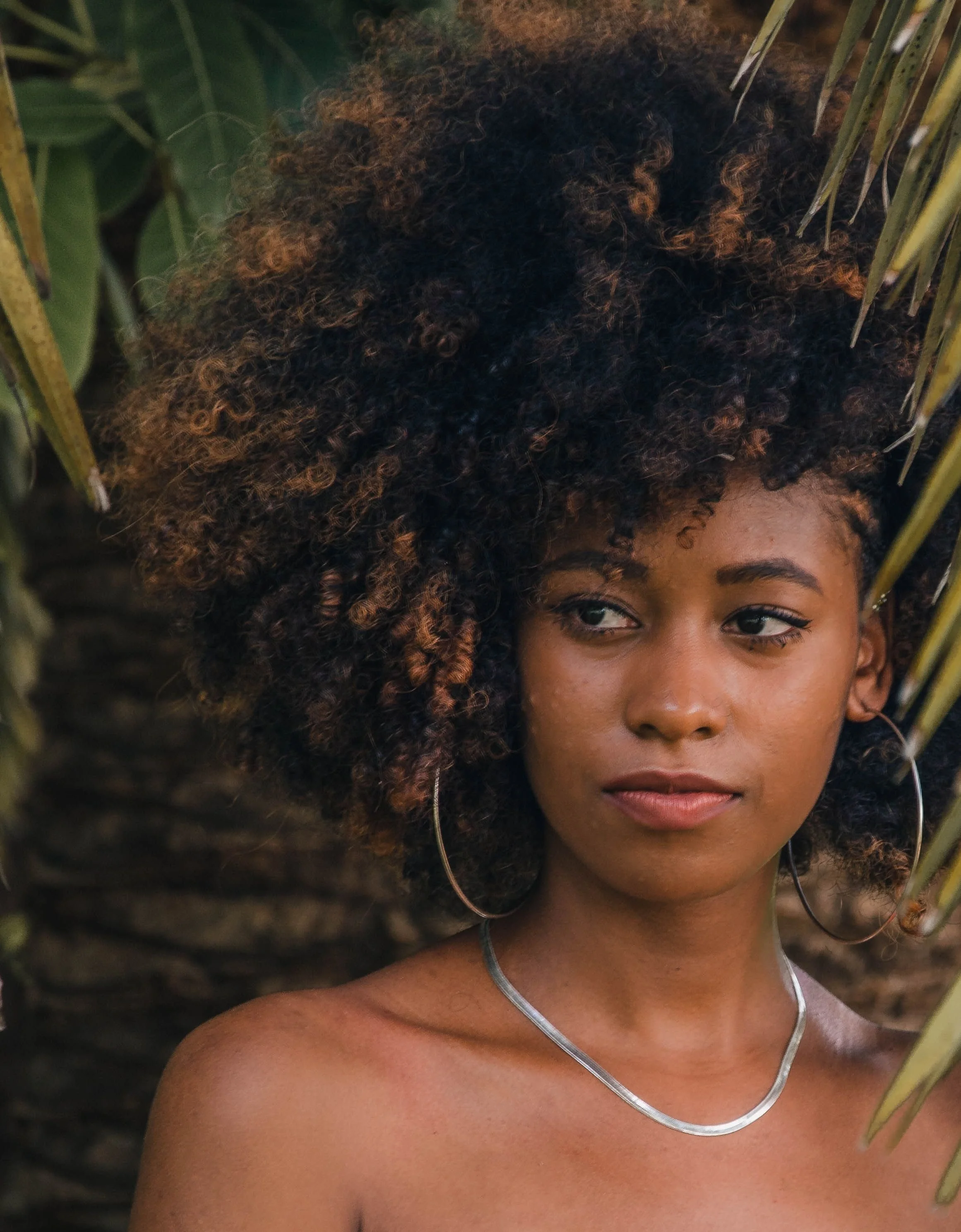 Close-up of a woman with curly hair, tan skin, wearing earrings and a necklace, against a background of green leaves and plants.