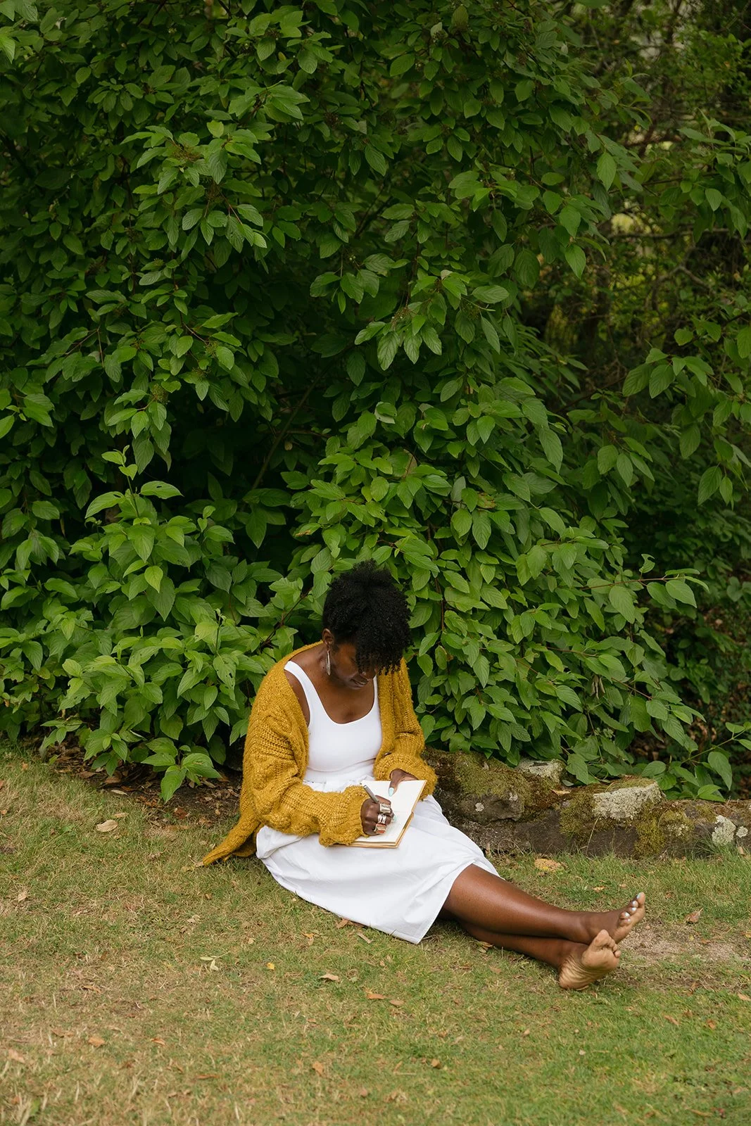 A woman in a white dress and mustard cardigan sitting on the grass, writing in a notebook, with a large green leafy bush behind her.