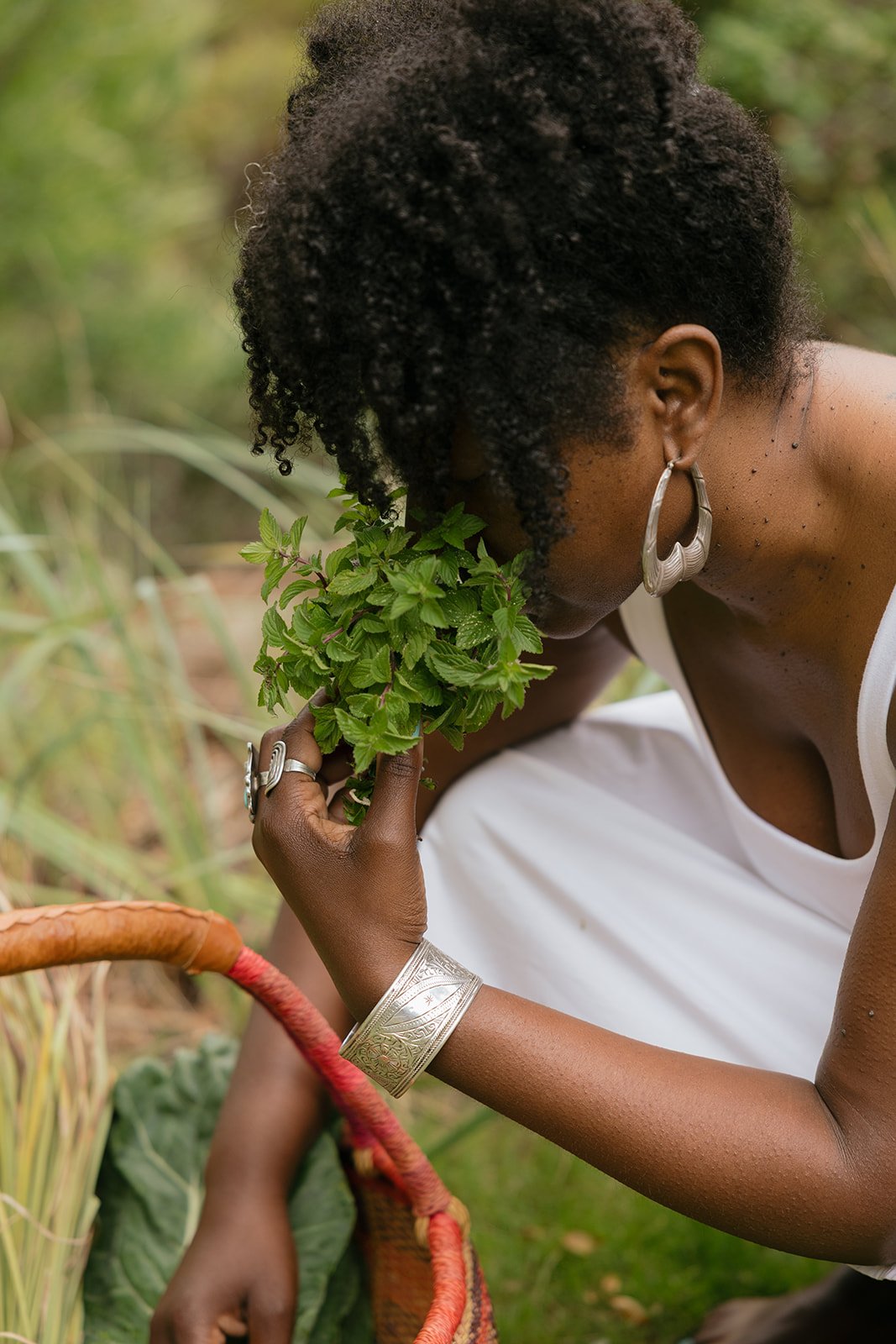 A woman with curly hair and large earrings, dressed in white, gently smelling a green plant while crouching outdoors.