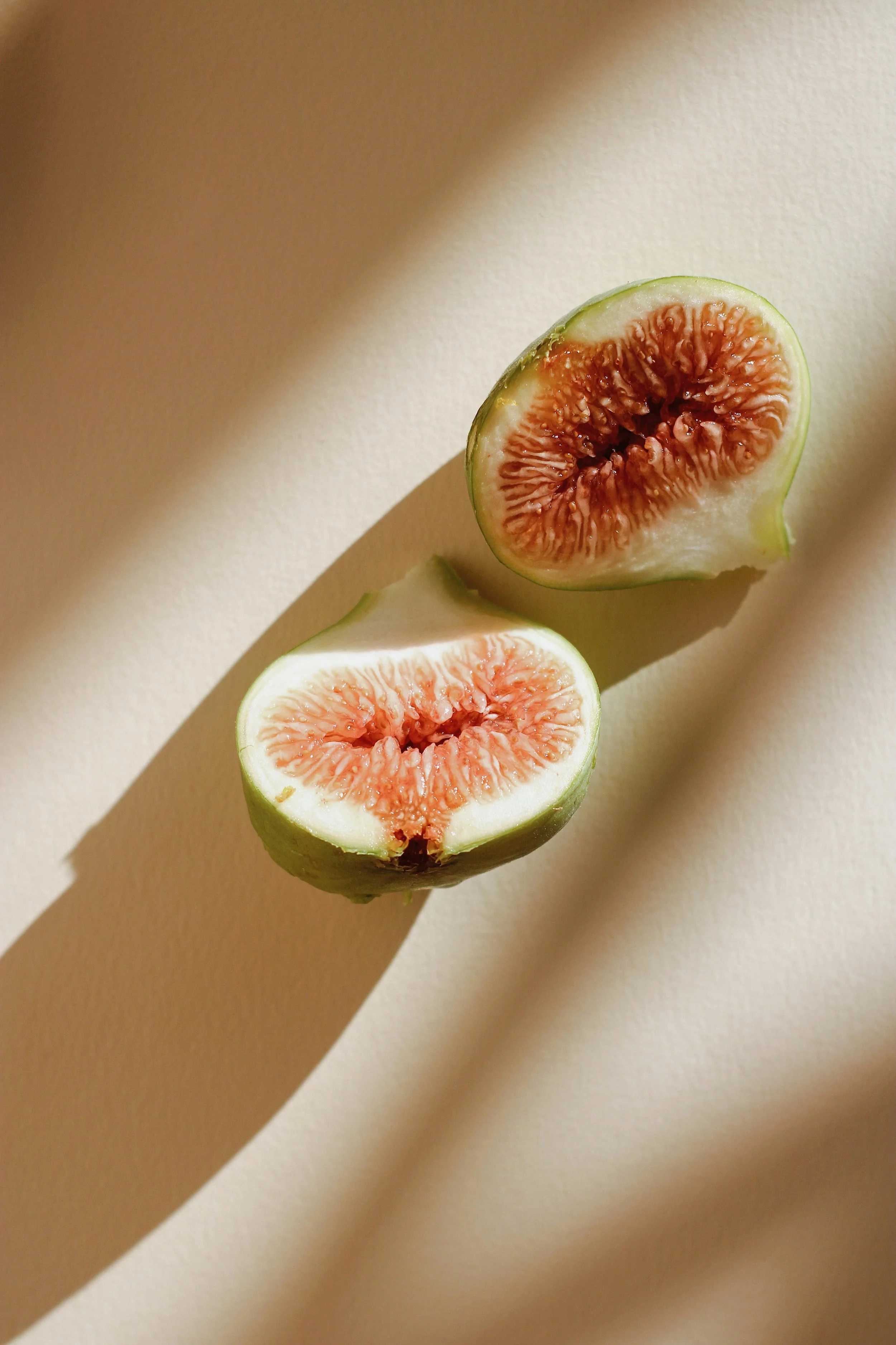 A sliced fig fruit with pink interior and green skin, casting a shadow on a light-colored surface.
