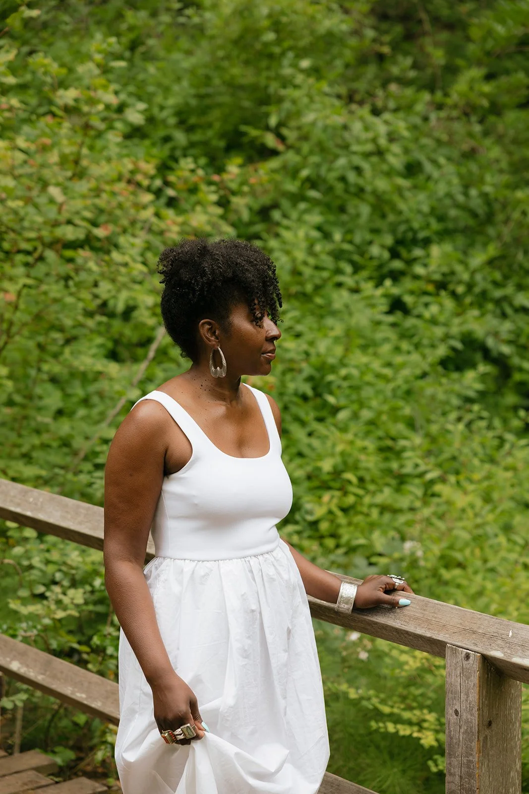 A woman dressed in white standing on a wooden bridge in a green outdoor setting.