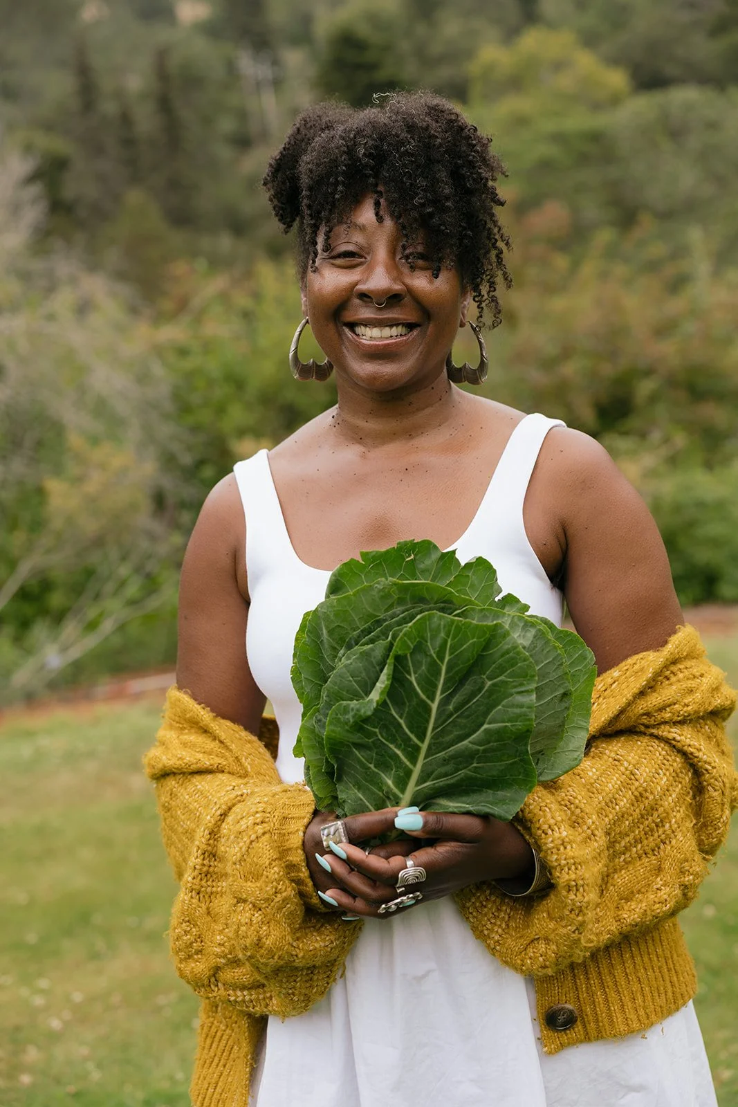 A woman smiling outdoors in a green garden holding a leafy vegetable, wearing a white dress, yellow cardigan, and large earrings.