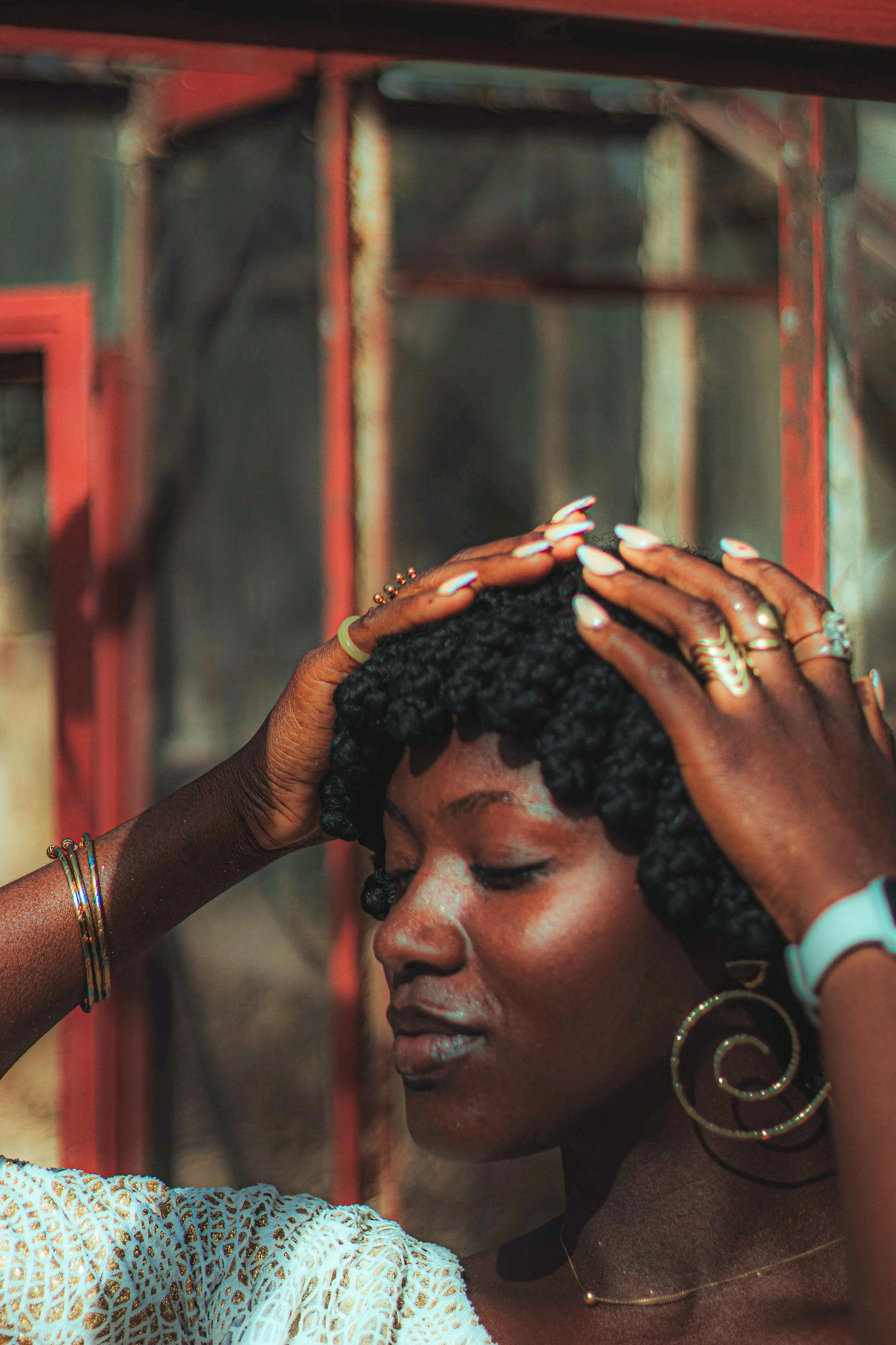 A woman with dark skin and braided hair, wearing large hoop earrings and multiple bracelets, has her eyes closed as two hands with painted nails rest on her head. The background shows a metal structure with red and black painted parts.