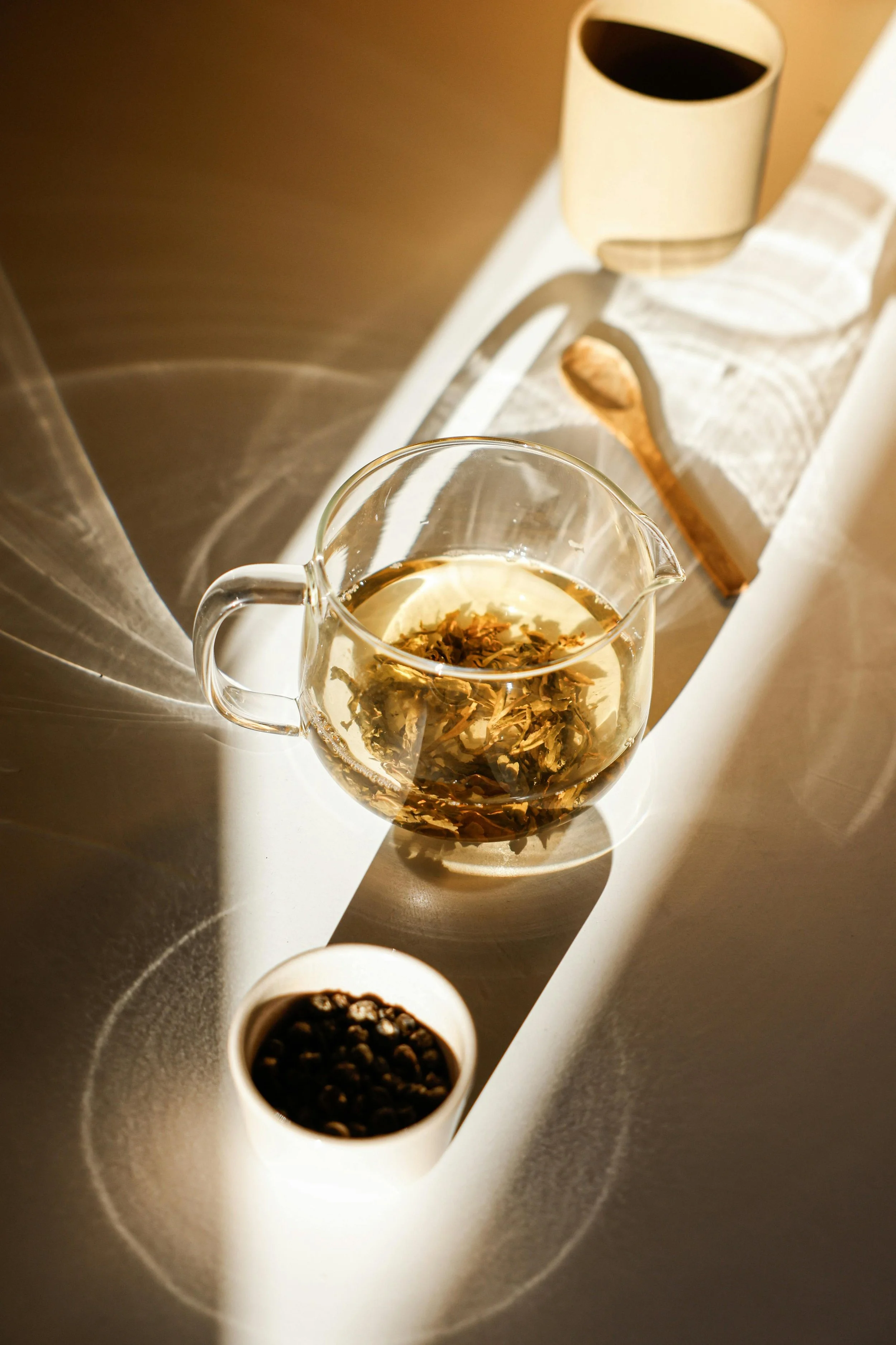 A glass pitcher of herbal tea with floating herbs, two white cups with black coffee, a small white bowl of peppercorns, and a wooden spoon on a white surface with sunlight casting shadows.