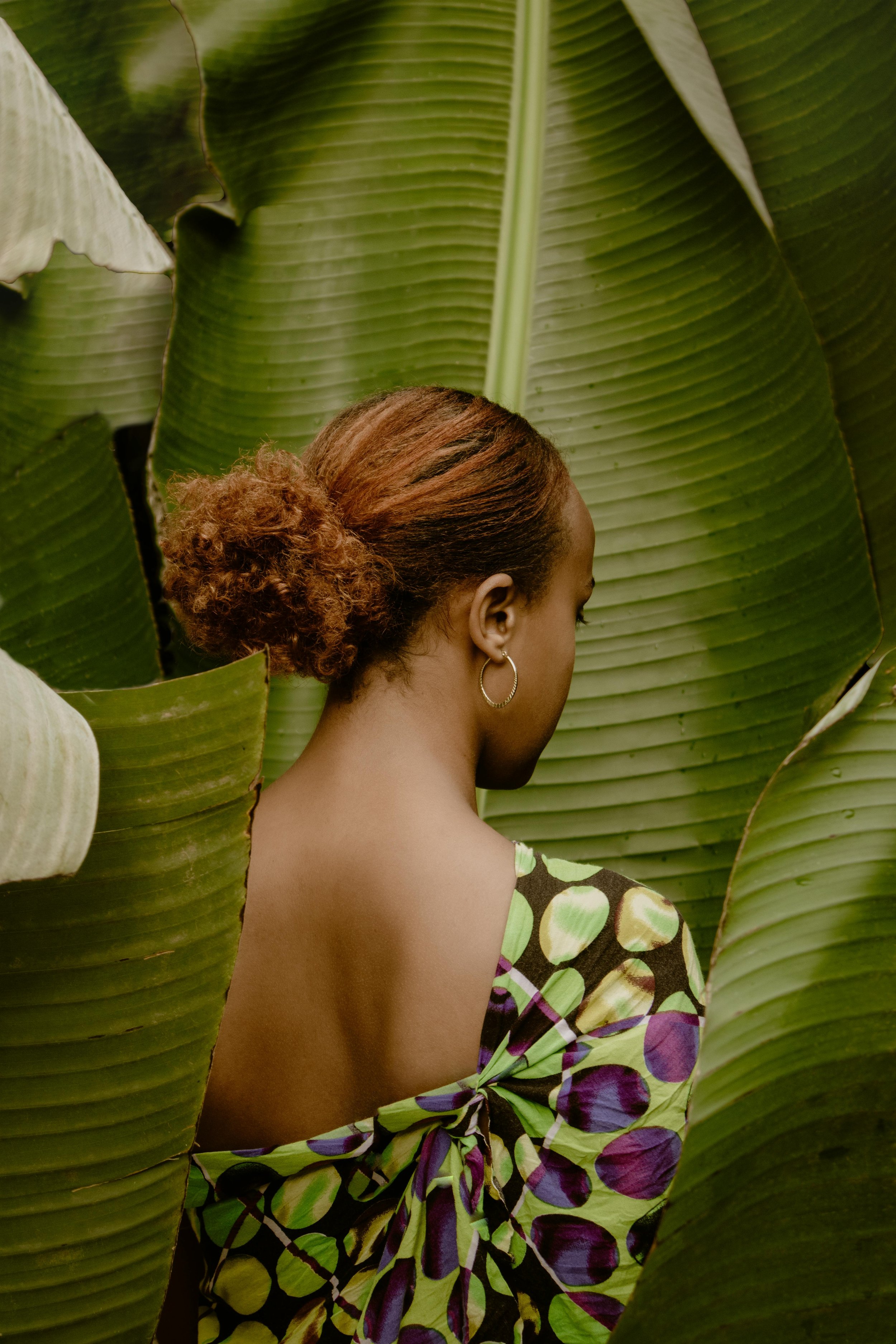 A woman with reddish-brown hair styled in a textured bun, wearing a patterned dress with purple, green, and black circles, standing among large green leaves.