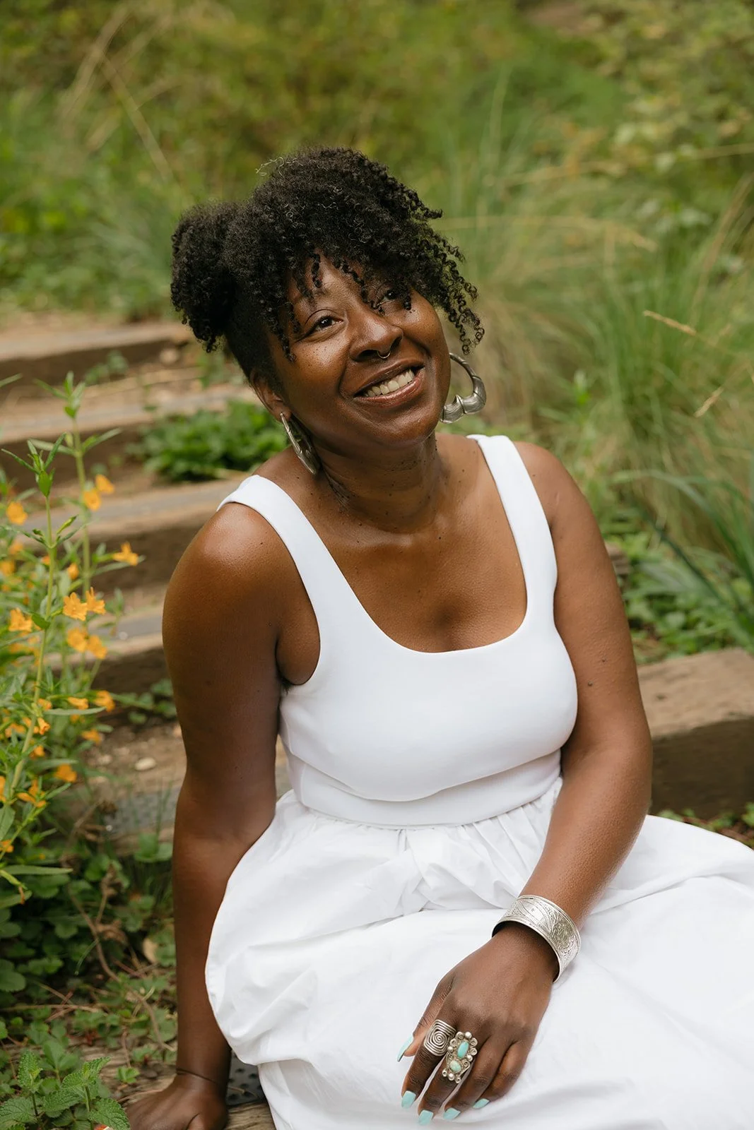 A woman in a white sleeveless dress sitting outdoors amidst green plants and flowers, smiling at the camera.
