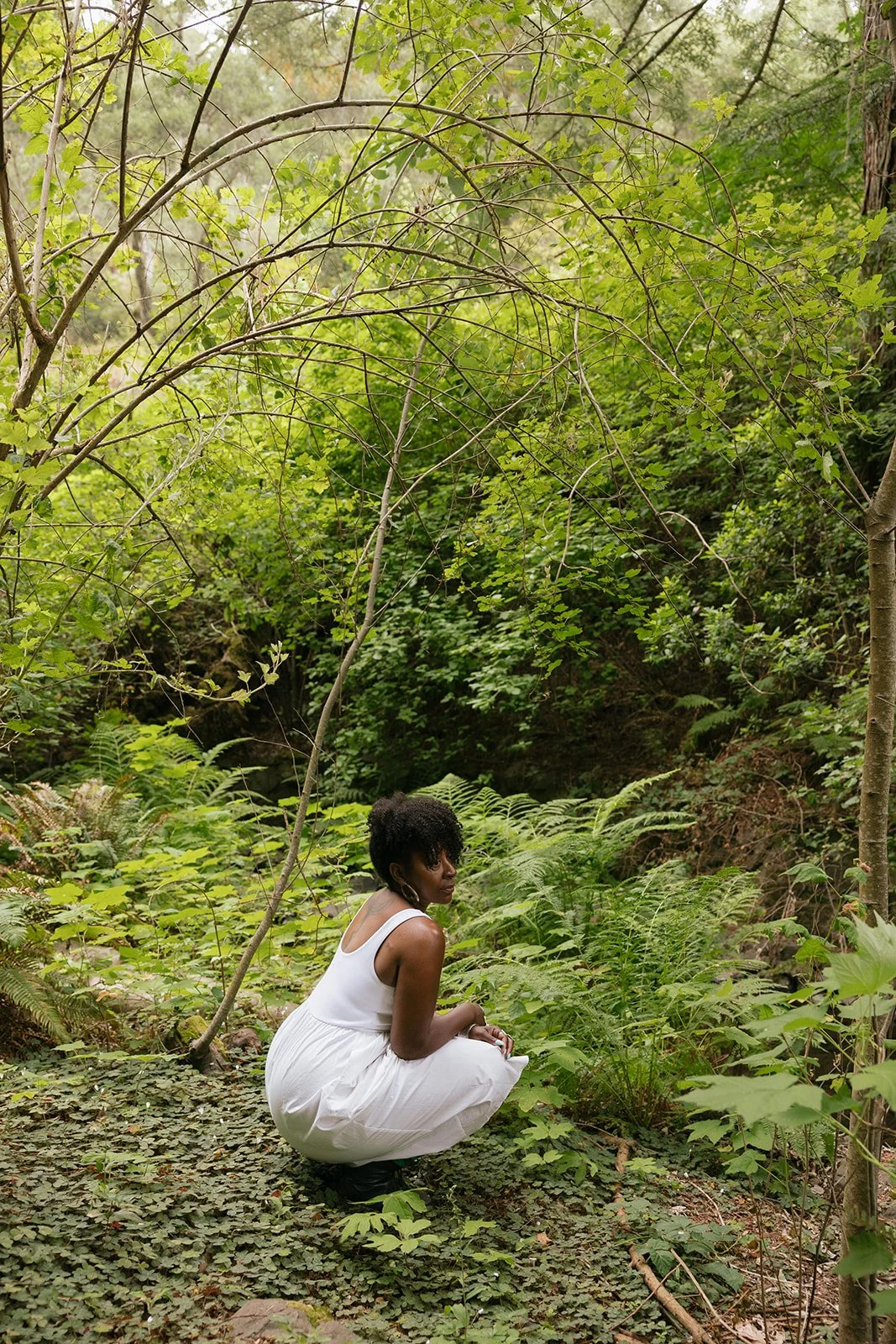 A woman in a white dress squatting in a lush green forest surrounded by ferns and foliage.