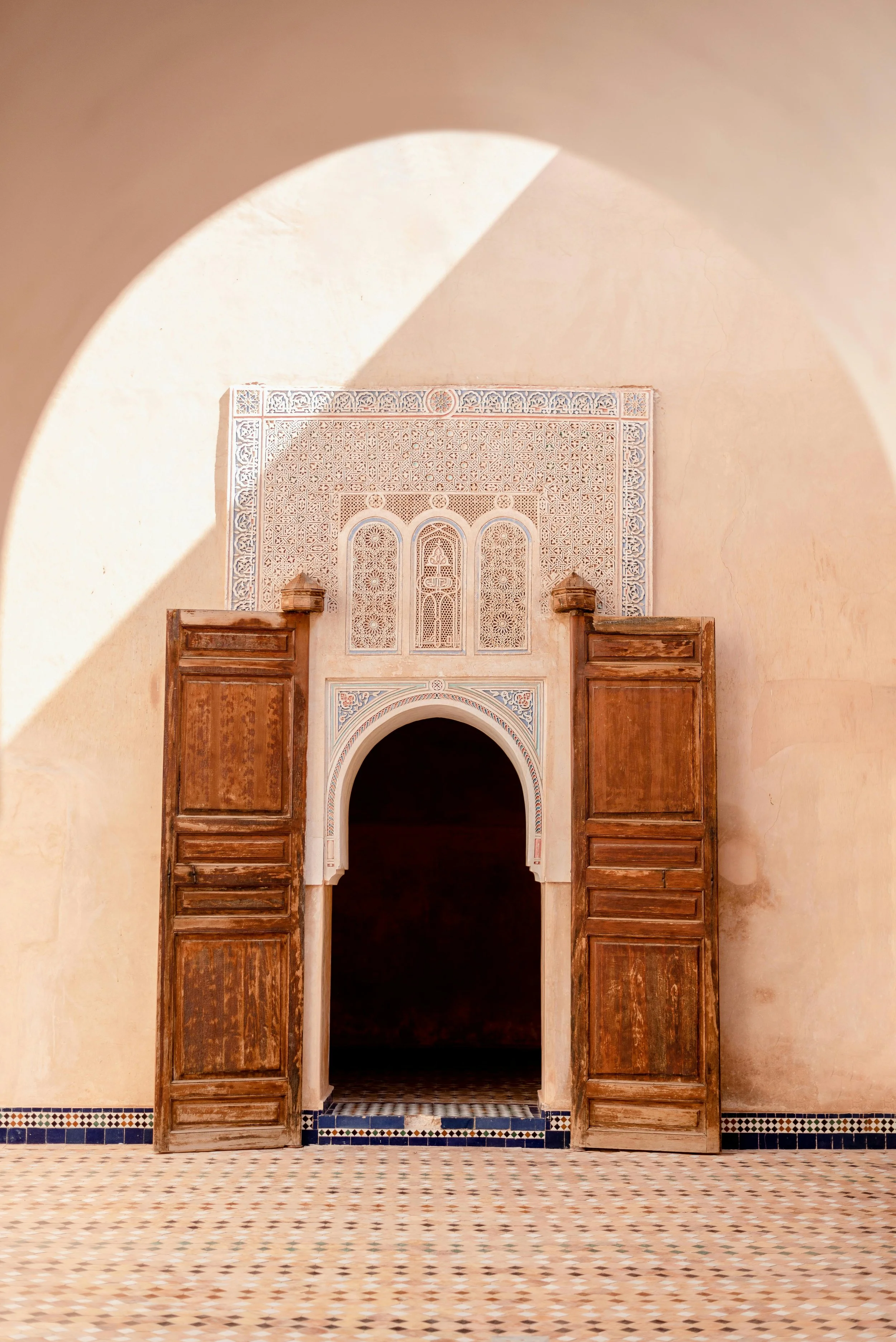 An ornate doorway with wooden shutters, intricate white plaster design above the arch, in a room with patterned tiled floors and walls with a warm, beige tone.