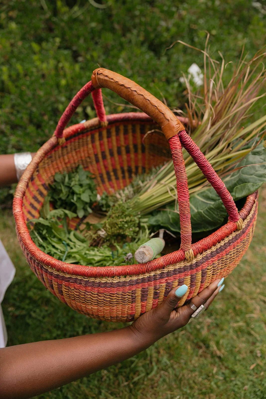 A colorful woven basket filled with various fresh vegetables and herbs, being held by a person with light blue nails.