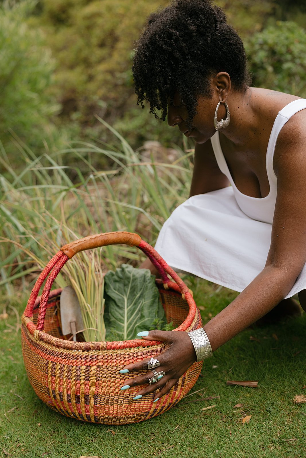 A woman in a white dress is picking vegetables, including yucca and leafy greens, from a garden and placing them into a basket.