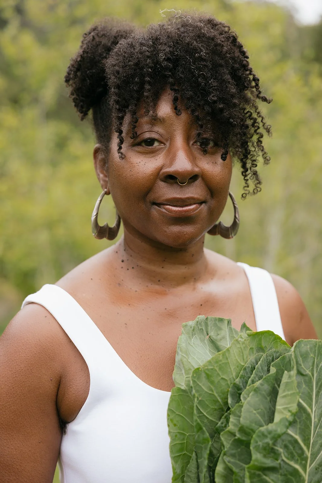 A woman with curly hair, wearing hoop earrings and a septum piercing, stands outdoors holding large green leafy vegetables, in a natural setting with blurred trees in the background.