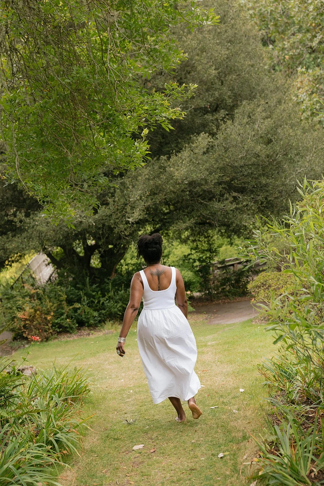 A woman in a white dress walking barefoot along a grassy garden path surrounded by green trees and plants.