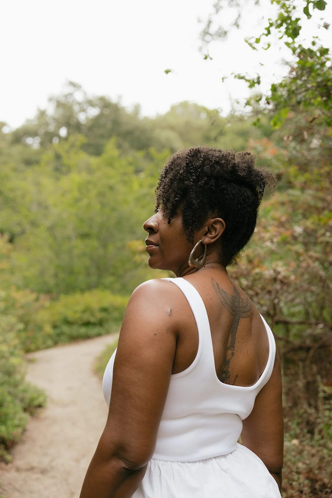 A woman standing outdoors on a dirt trail surrounded by green trees, wearing a white sleeveless top and earrings, with short curly hair and a tattoo on her back.