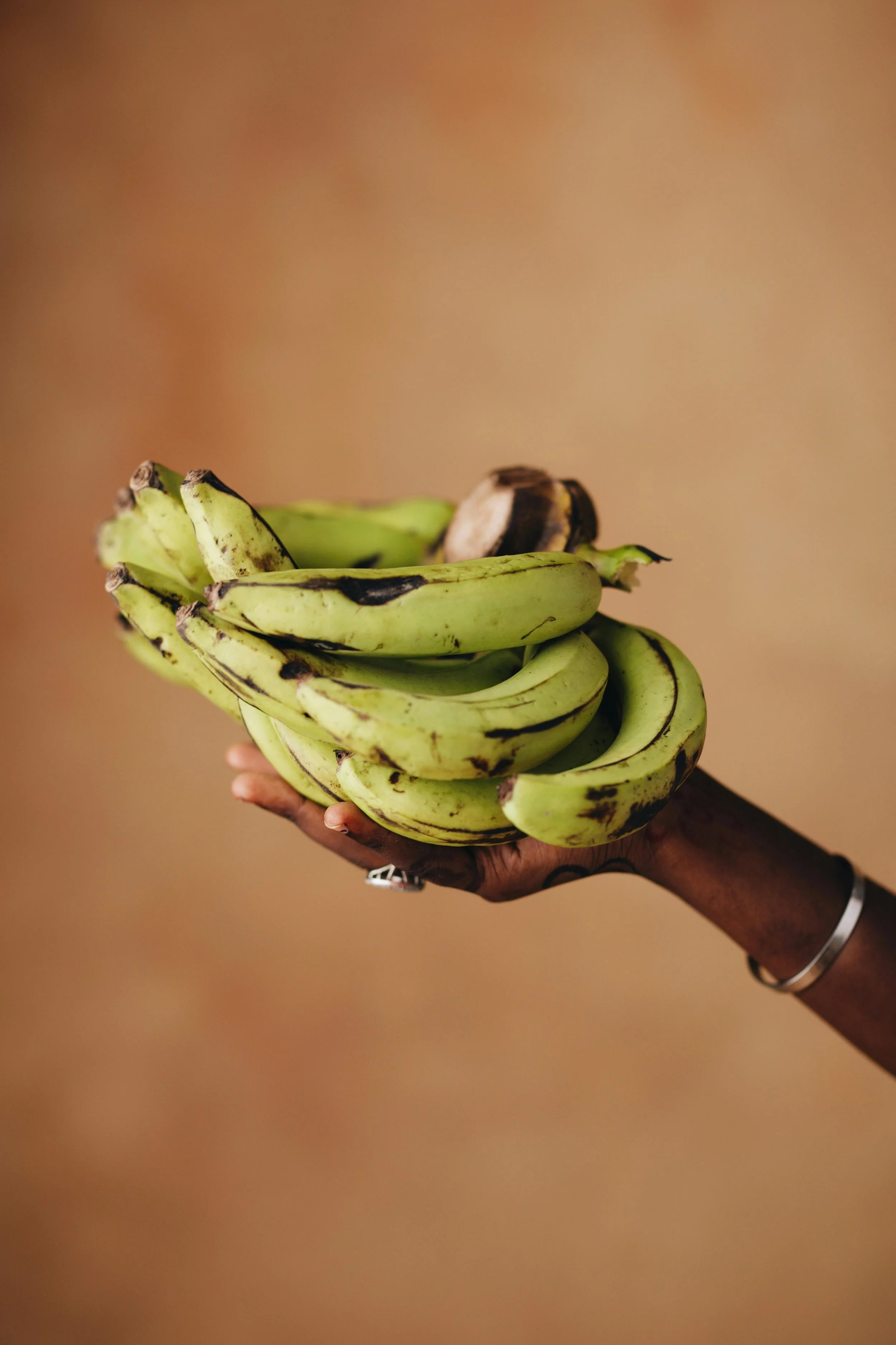A person with dark skin holding a bunch of green bananas with some brown spots, against a plain background.