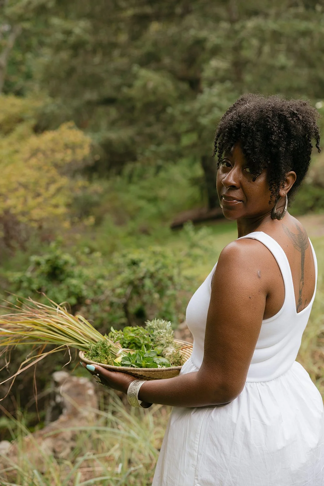 Woman in a white dress holding a basket of herbs outdoors, with lush green trees and foliage in the background.