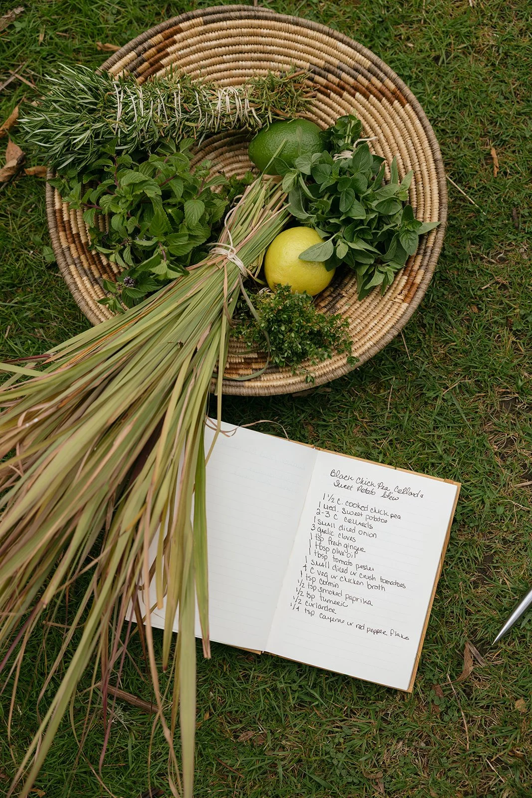 A woven basket containing fresh herbs, a lime, a lemon, and a bundle of dried herbs, placed on green grass with an open handwritten recipe book nearby.