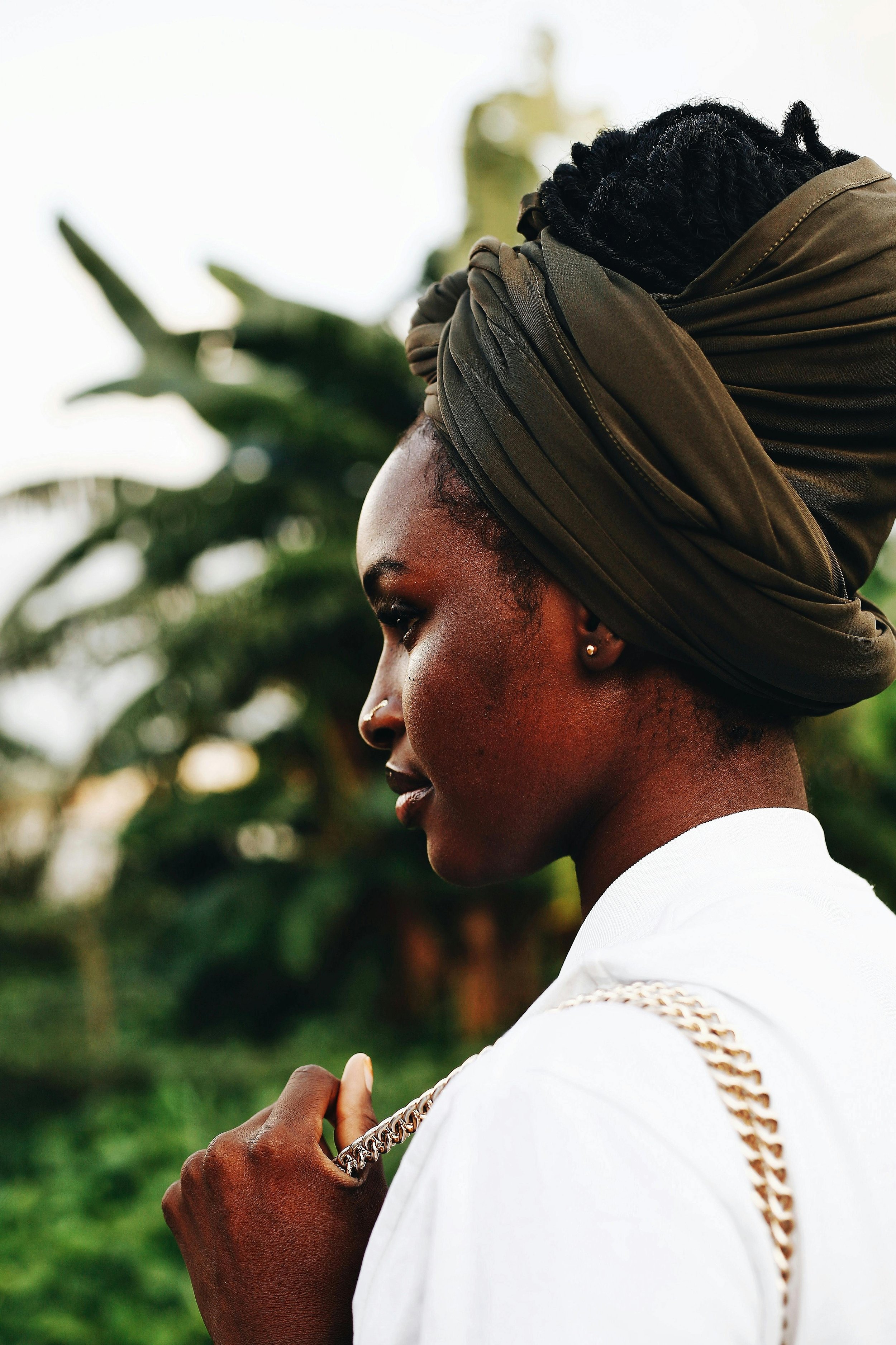 A Black woman with short hair, wearing a head wrap and a white shirt, holding a chain necklace in her hand, outdoors with blurred greenery in the background.