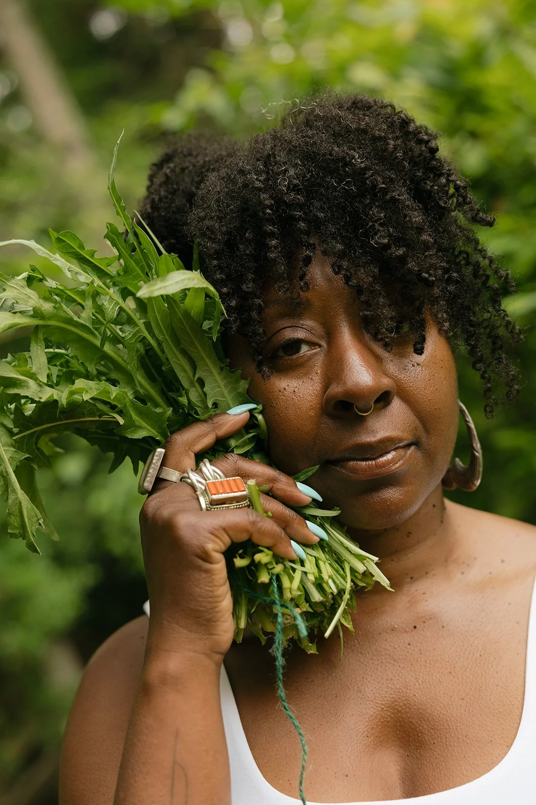 A woman with curly black hair and hoop earrings holding a bunch of green leafy plants.