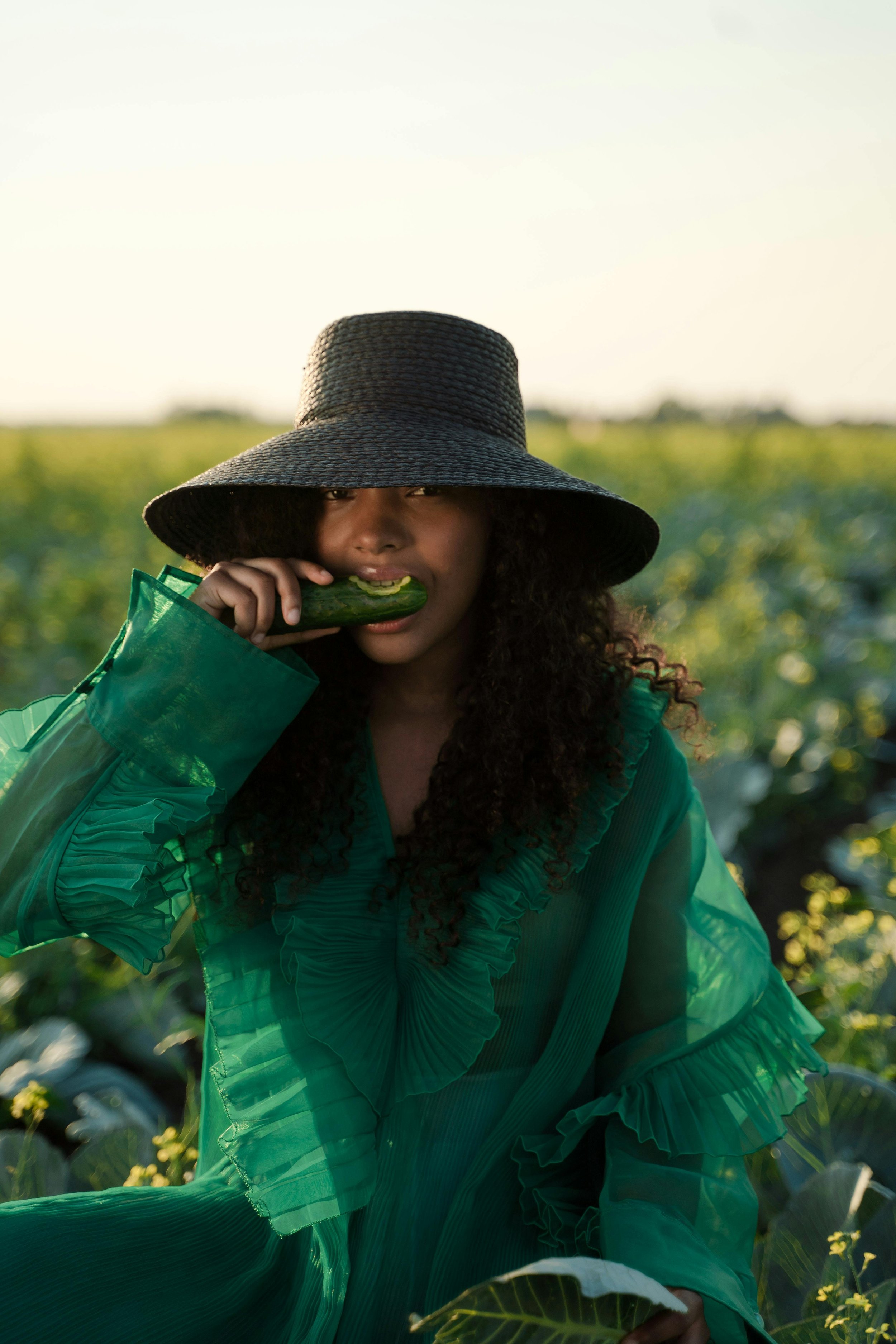 Woman in green ruffled dress and large floppy hat biting a cucumber in a lush green field during daylight.