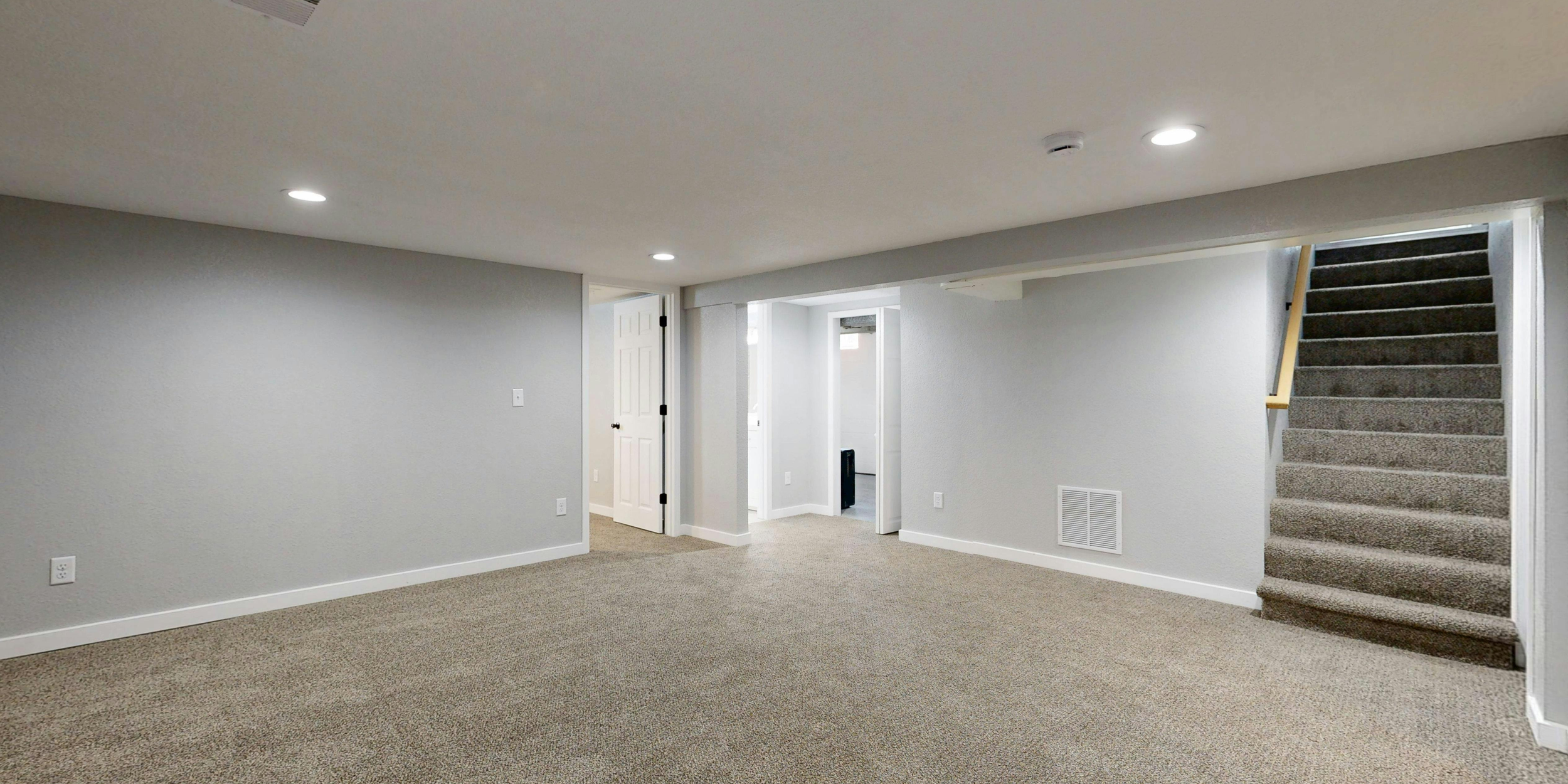 Empty basement room with beige carpet, white walls, and recessed lighting; staircase with carpeted steps and wooden handrail, open doorways leading to other rooms.