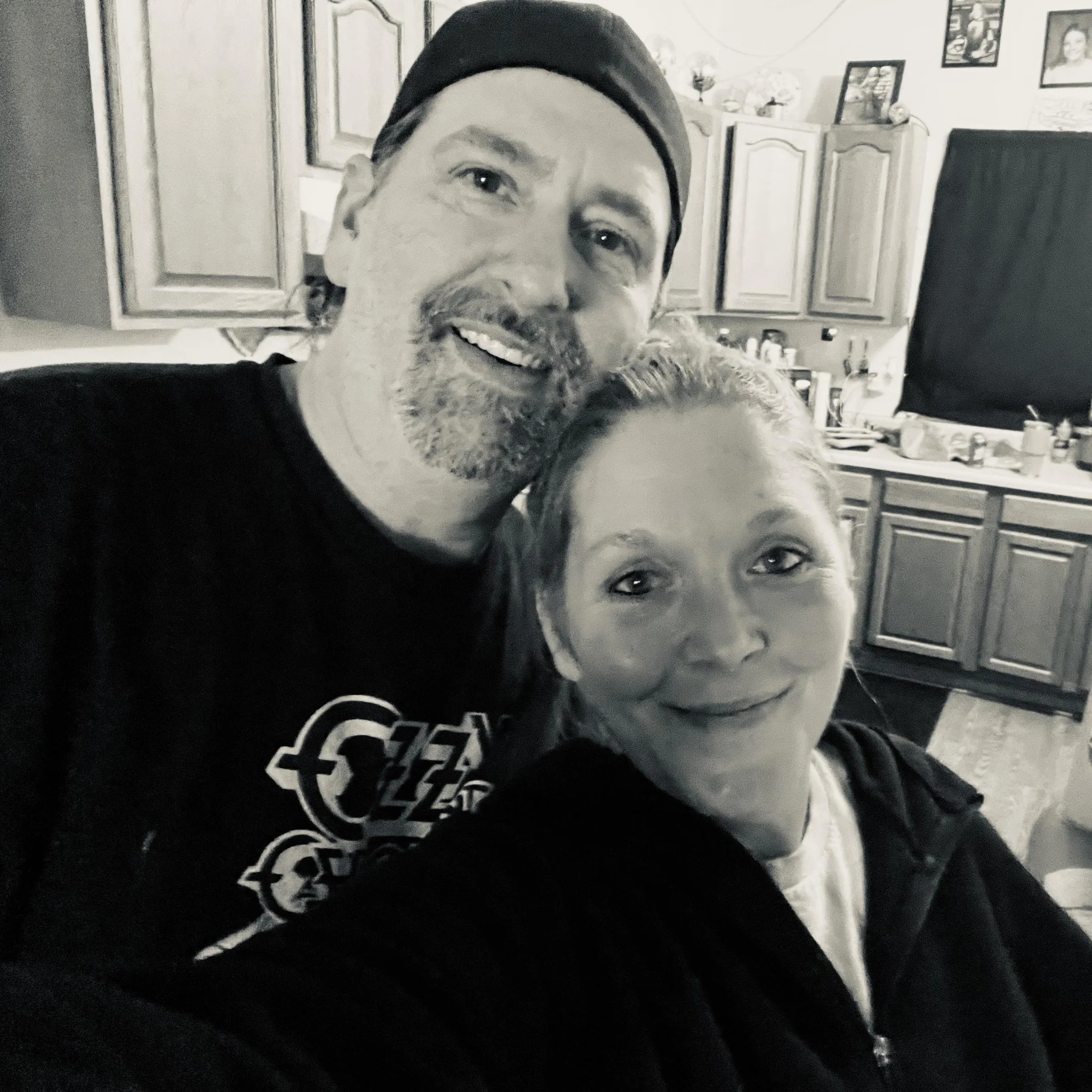 A man with a beard and mustache wearing a beanie, smiling while holding a dog. The background shows kitchen cabinets, framed photos, and kitchen items.