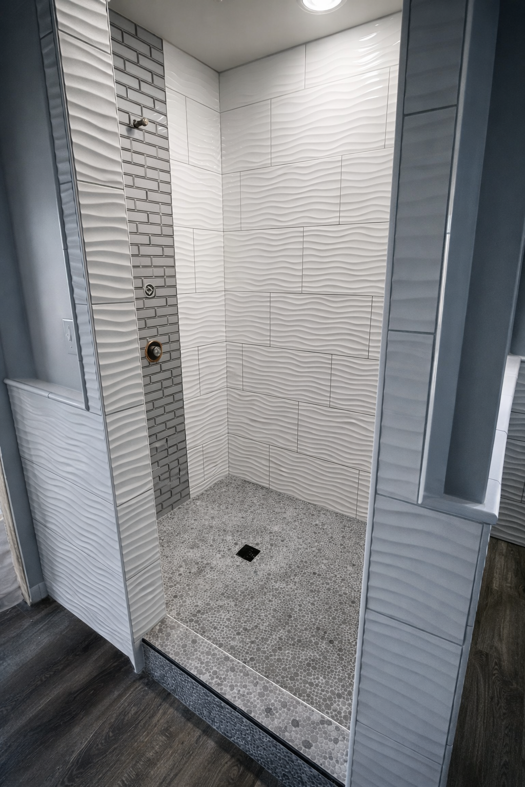 View of a modern shower area with white textured wall tiles, and a small dark grey tile accent strip. The shower floor features small, irregular grey stones, and there is a drain at the center. The shower is enclosed by partial framing with a textured surface.