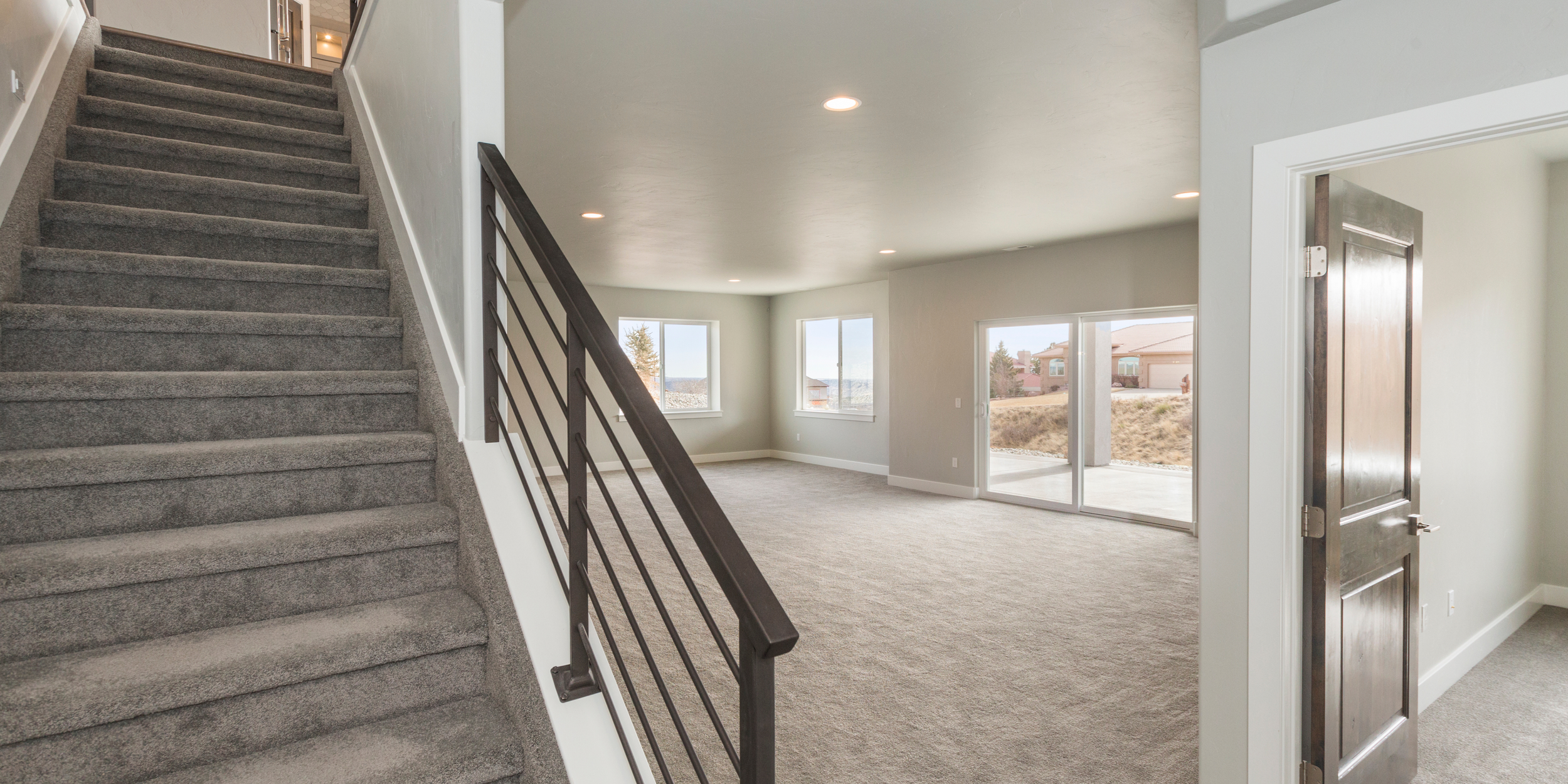 An empty modern living room with large windows, sliding glass door, carpeted floor, white walls, and recessed ceiling lights, view from staircase.