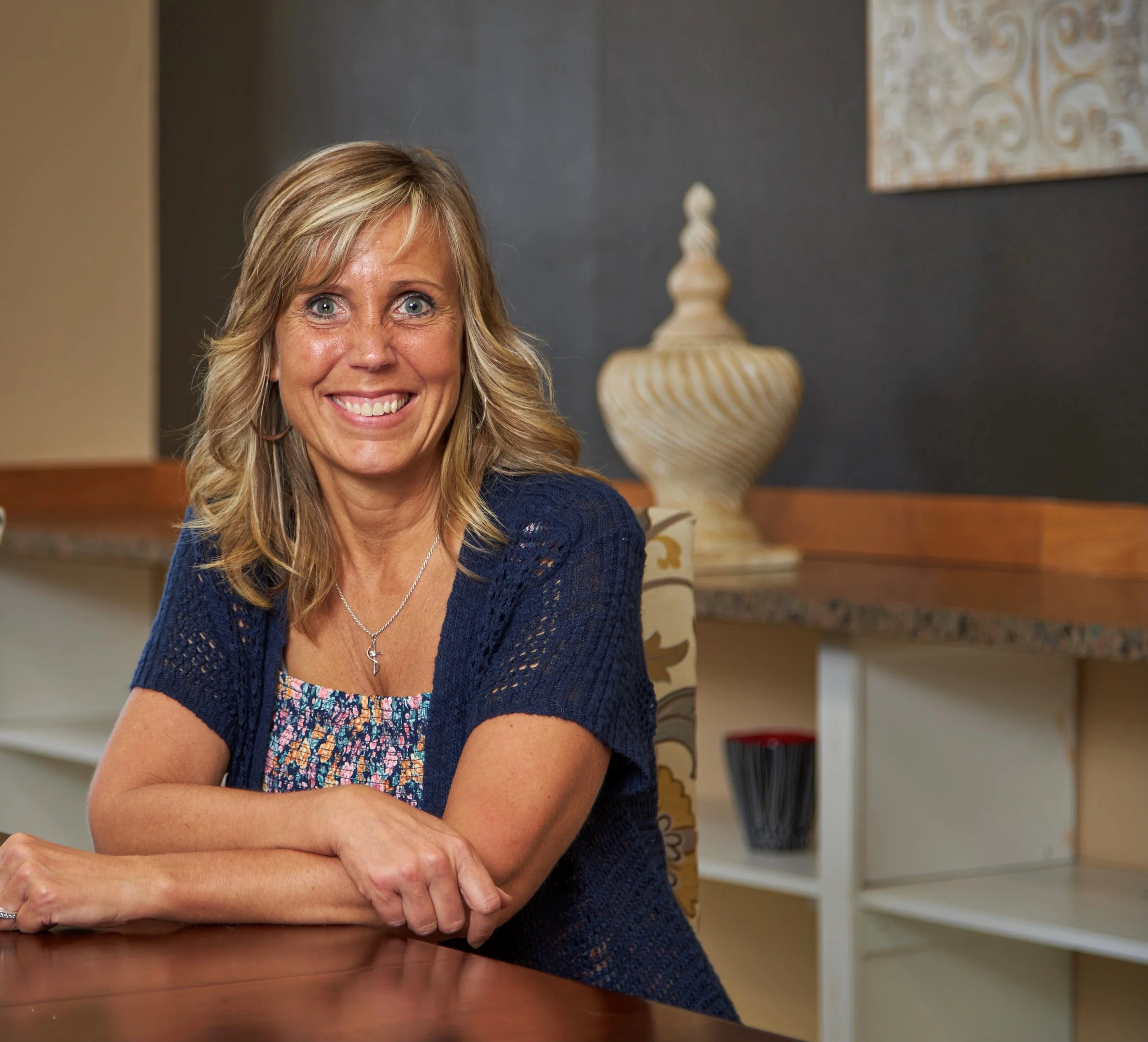 Close-up of a smiling woman with blonde hair, wearing a blue cardigan and a patterned top, sitting at a wooden table in a room with dark gray walls, a decorative ceramic vase on a shelf, and a framed artwork on the wall.