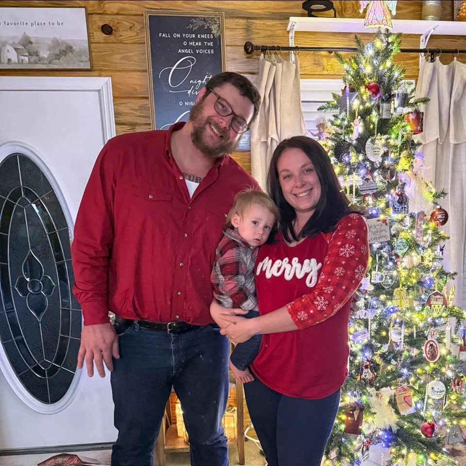 A group of three people, a man, a woman, and a young boy, poses in front of a decorated Christmas tree inside a cozy home. The man and woman are smiling, while the boy looks slightly serious. The woman is wearing a red sweater with the word 'Merry,' and the man is wearing a red shirt. The Christmas tree is adorned with lights, ornaments, and ribbons, creating a festive atmosphere.