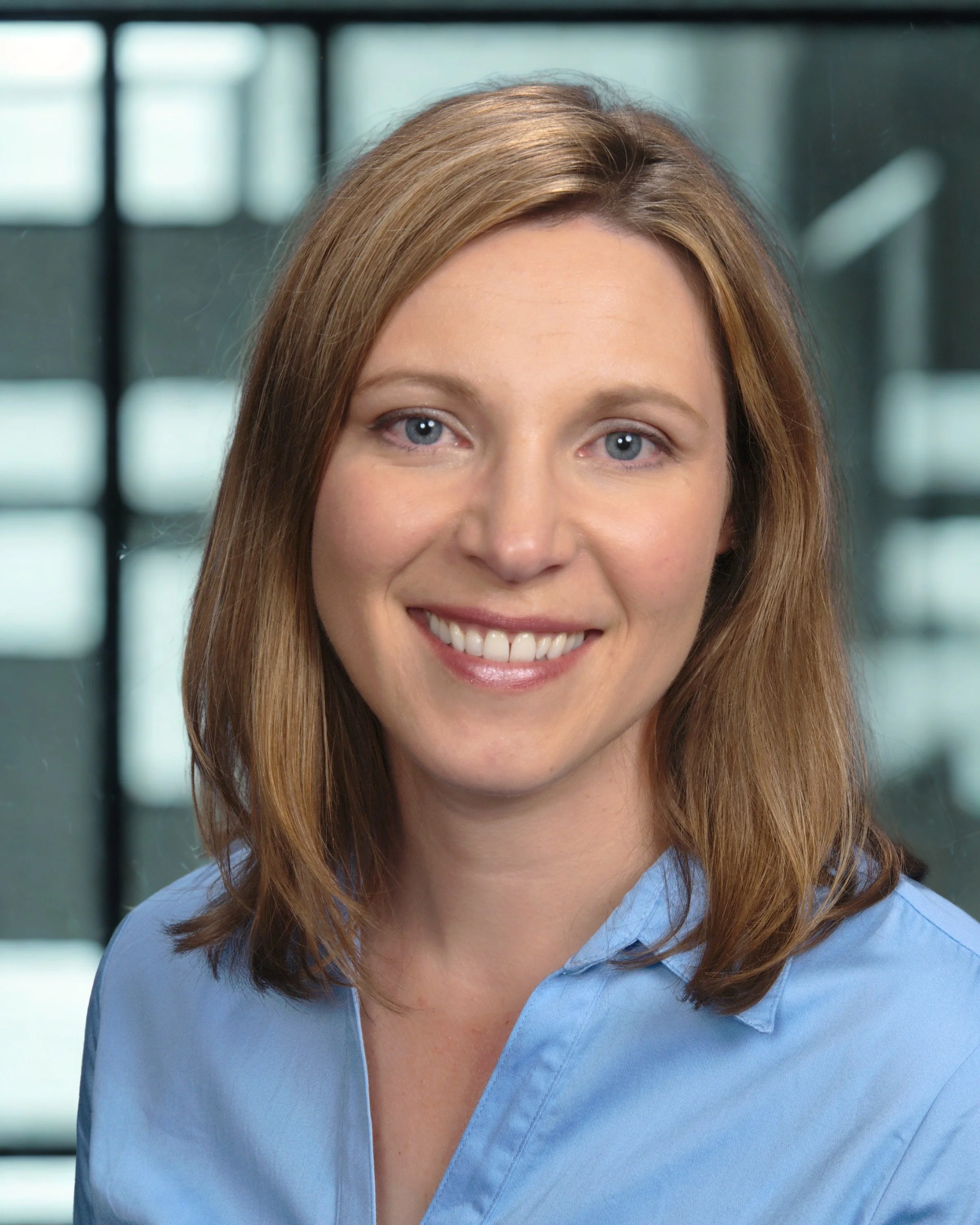 Close-up of a woman smiling, wearing a light blue shirt, with an office background.