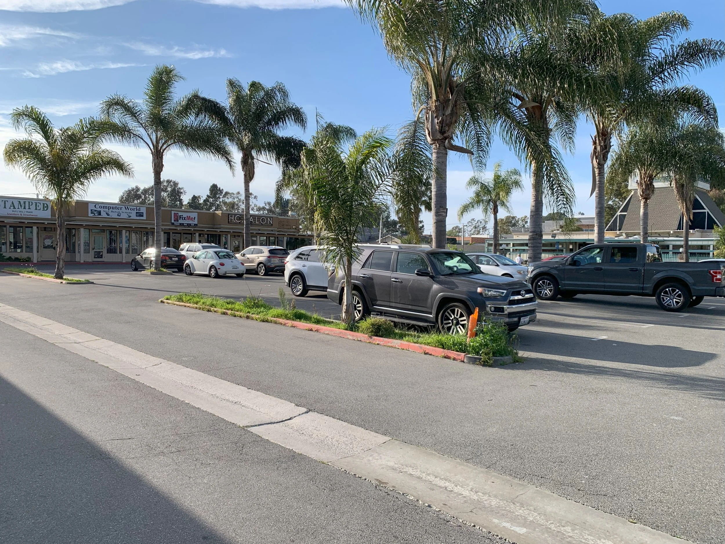 Parking lot with trees, buildings, and parked cars under a blue sky.