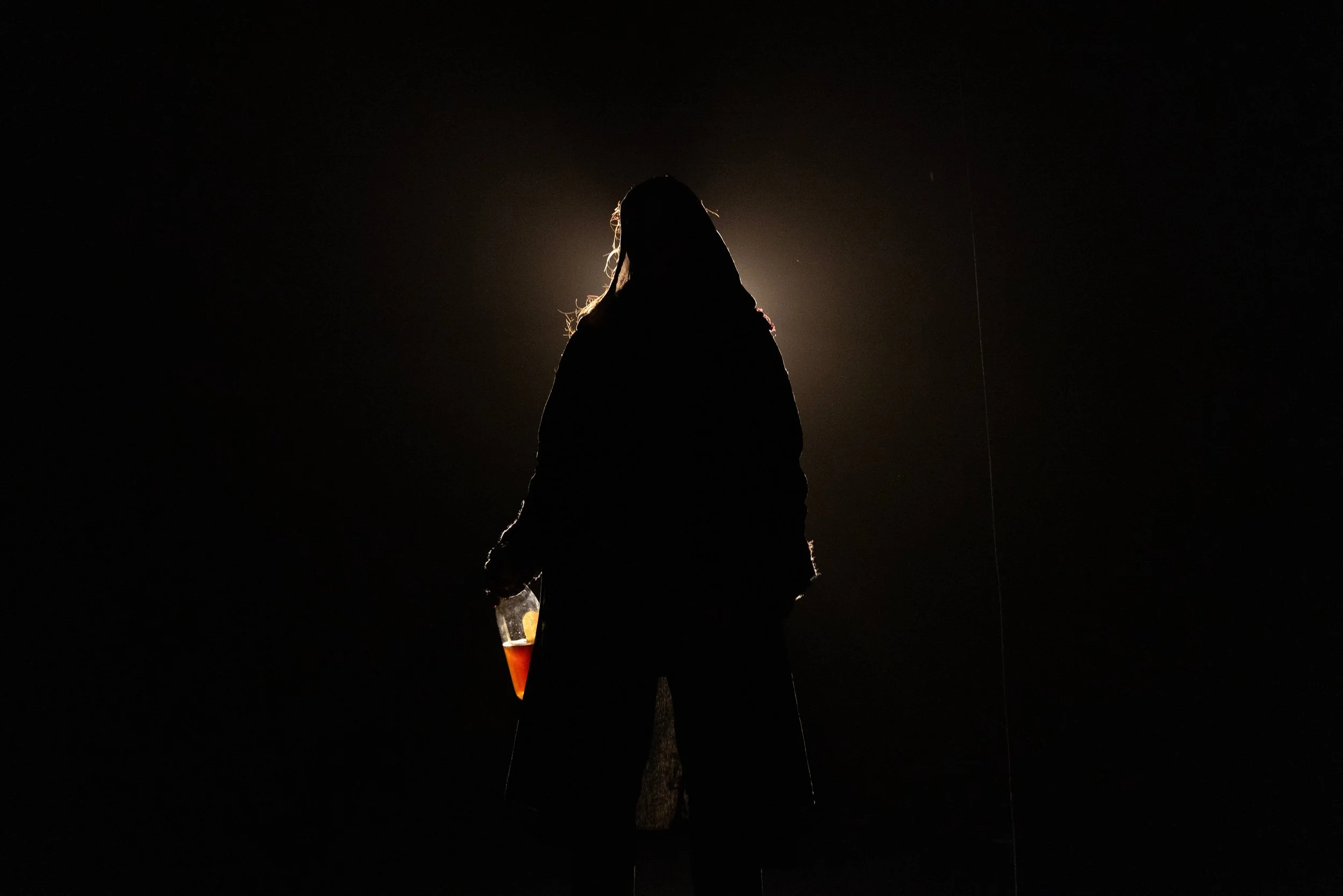 A silhouette of a person standing outdoors at night, backlit by a bright light, holding a drink in a glass, with long hair, possibly a woman, and standing near a tree.