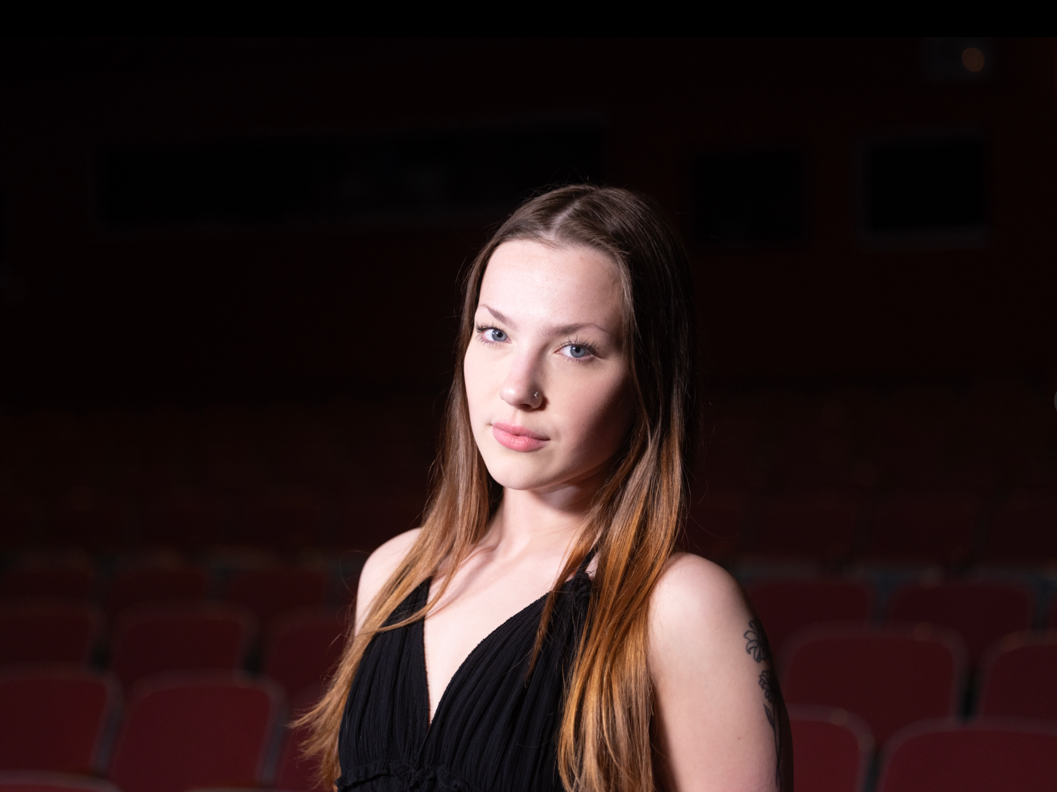 A young woman with light skin, long brown hair, blue eyes, and a nose piercing looking at the camera. She is wearing a black sleeveless top. The background appears to be a theater or auditorium with dark lighting and red seats.