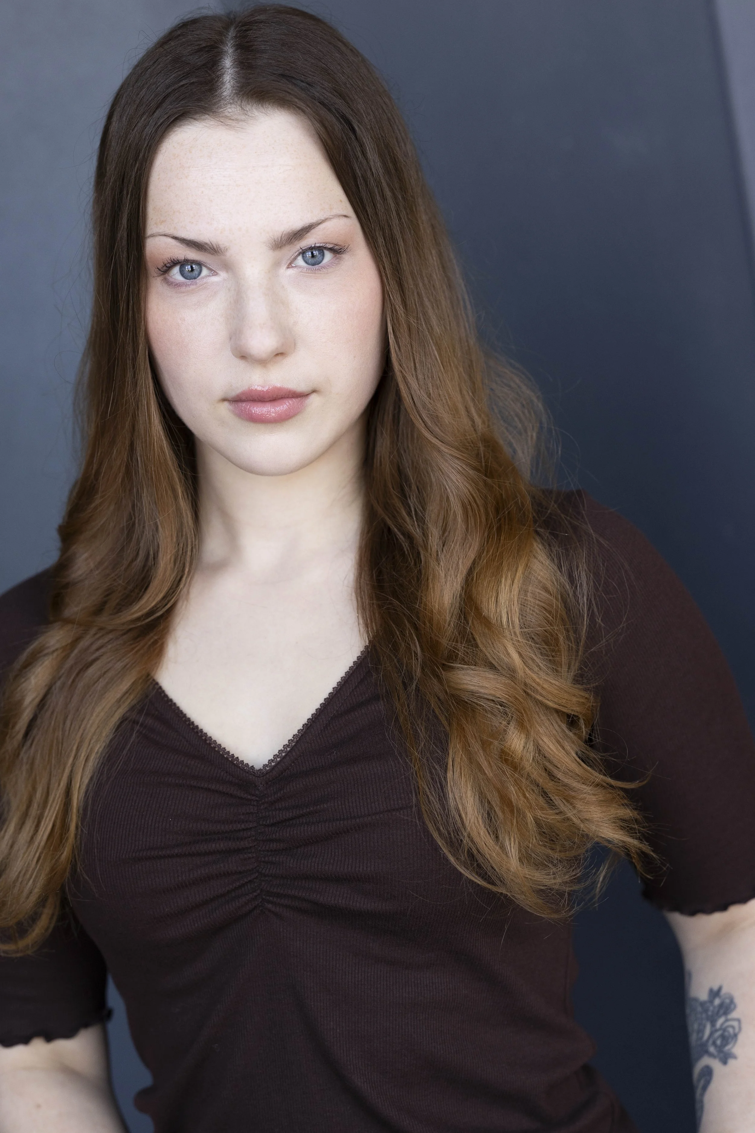 Close-up of a young woman with long, wavy red hair, blue eyes, and light skin, wearing a dark brown shirt, against a gray background.