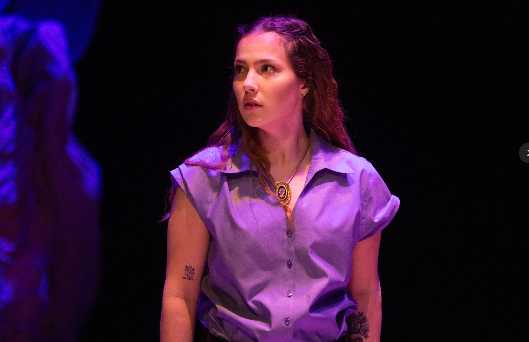 A woman with long reddish-brown hair and a tattoo on her forearm, wearing a light purple shirt with rolled-up sleeves and a decorative necklace, looking off to the side with a serious expression, under stage lighting.