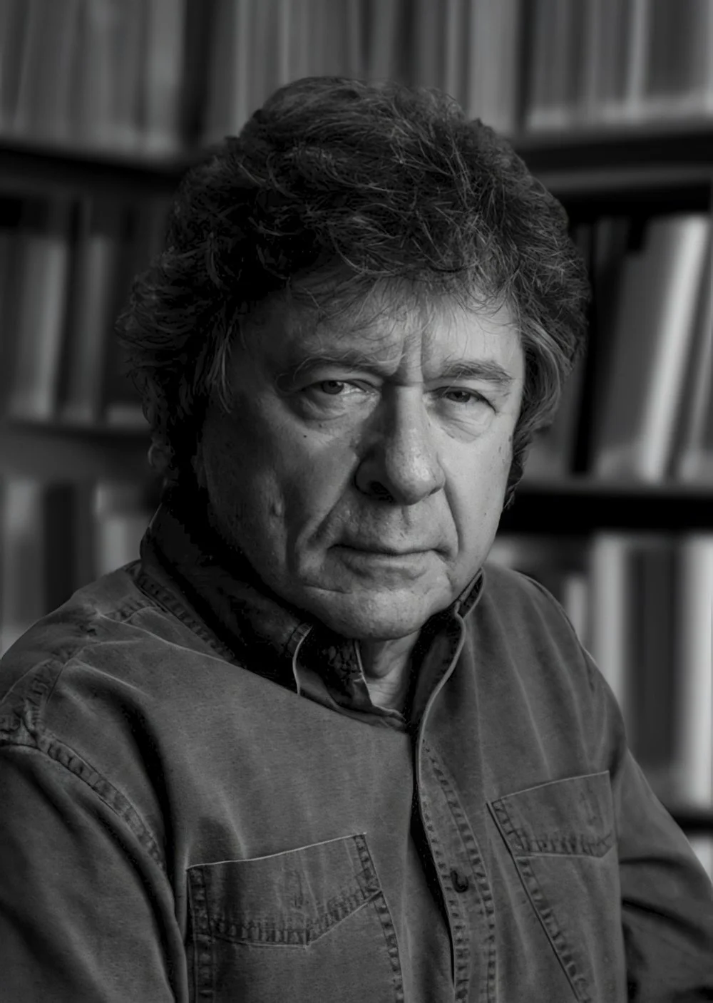 Black and white portrait of a middle-aged man with wavy hair, wearing a denim shirt, in front of a bookshelf filled with books.