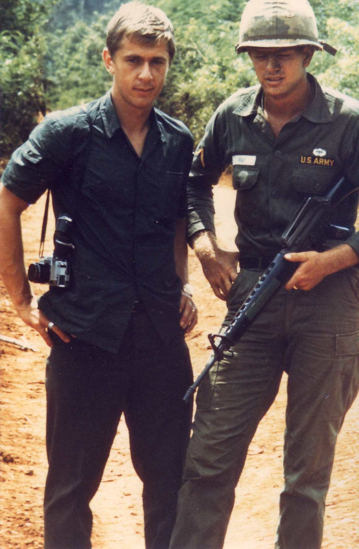 A young man and a U.S. Army soldier stand outdoors on a dirt path surrounded by greenery. The young man, Martyn Burke wears a military shirt and has a camera hanging around his neck. The soldier wears a military uniform and helmet, holding a rifle.