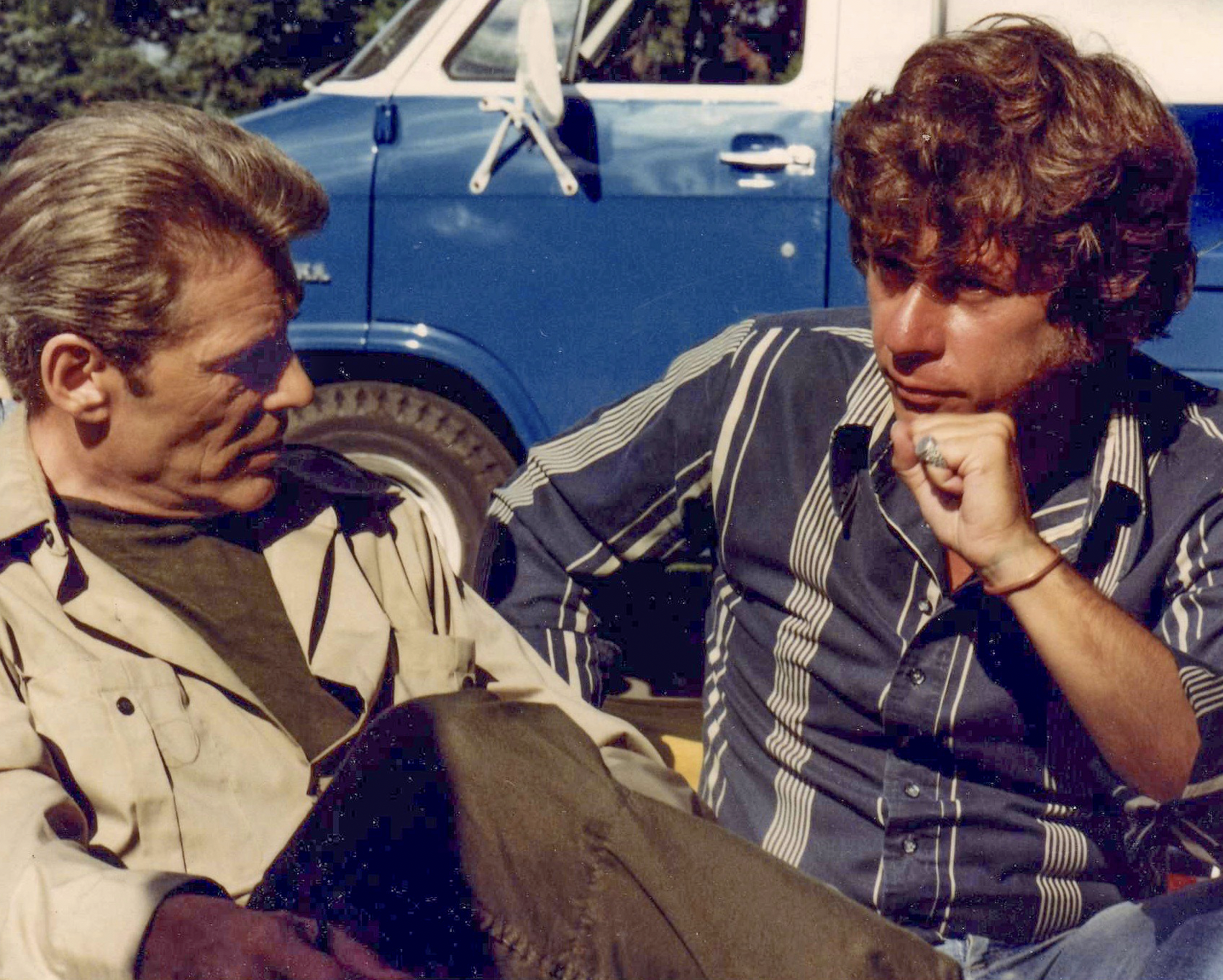 Two men sitting outdoors on a sunny day, having a conversation; a blue vintage vehicle is parked behind them. The man on the left is actor Peter O'Toole and the man on the right is author and filmmaker Martyn Burke.