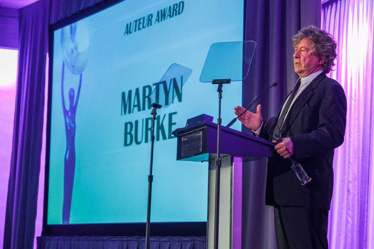A man in a suit standing at a podium giving a speech at an awards ceremony with a large screen behind him displaying text that reads 'Auteur Award' and 'Martyn Burke'.