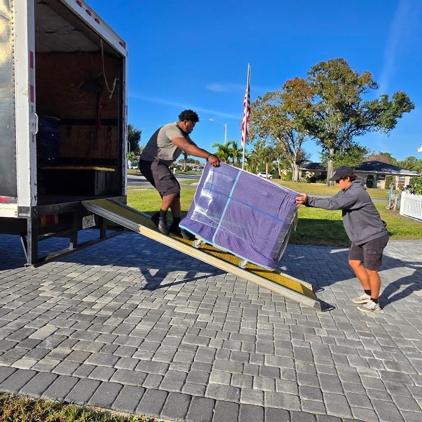 Two men are unloading a large wrapped piece of furniture from a moving truck onto a paved driveway outdoors with trees, houses, and an American flag in the background.