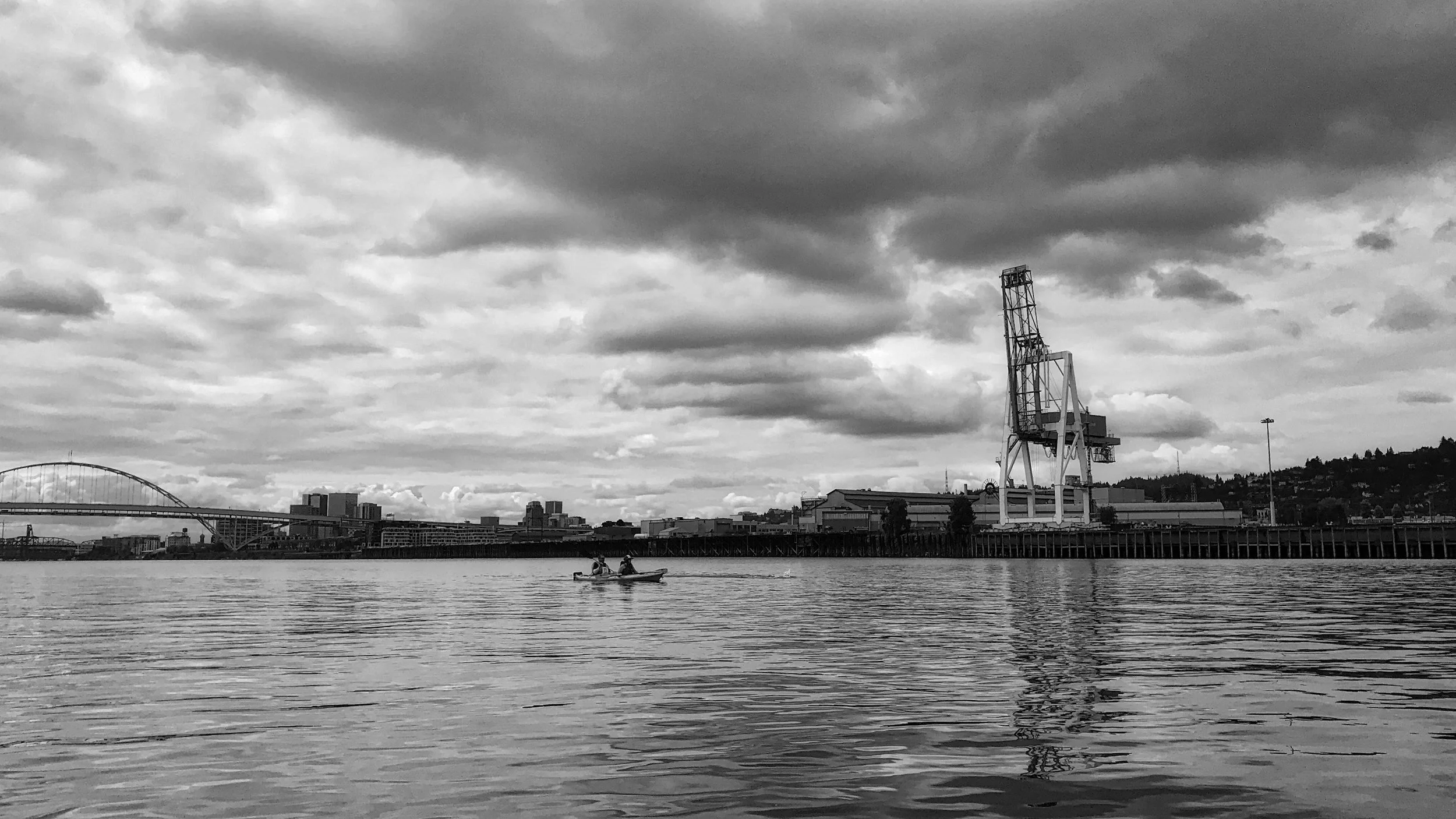 A grayscale landscape image of a two-person kayak on the the Willamette River looking upriver towards the Freemont Bridge