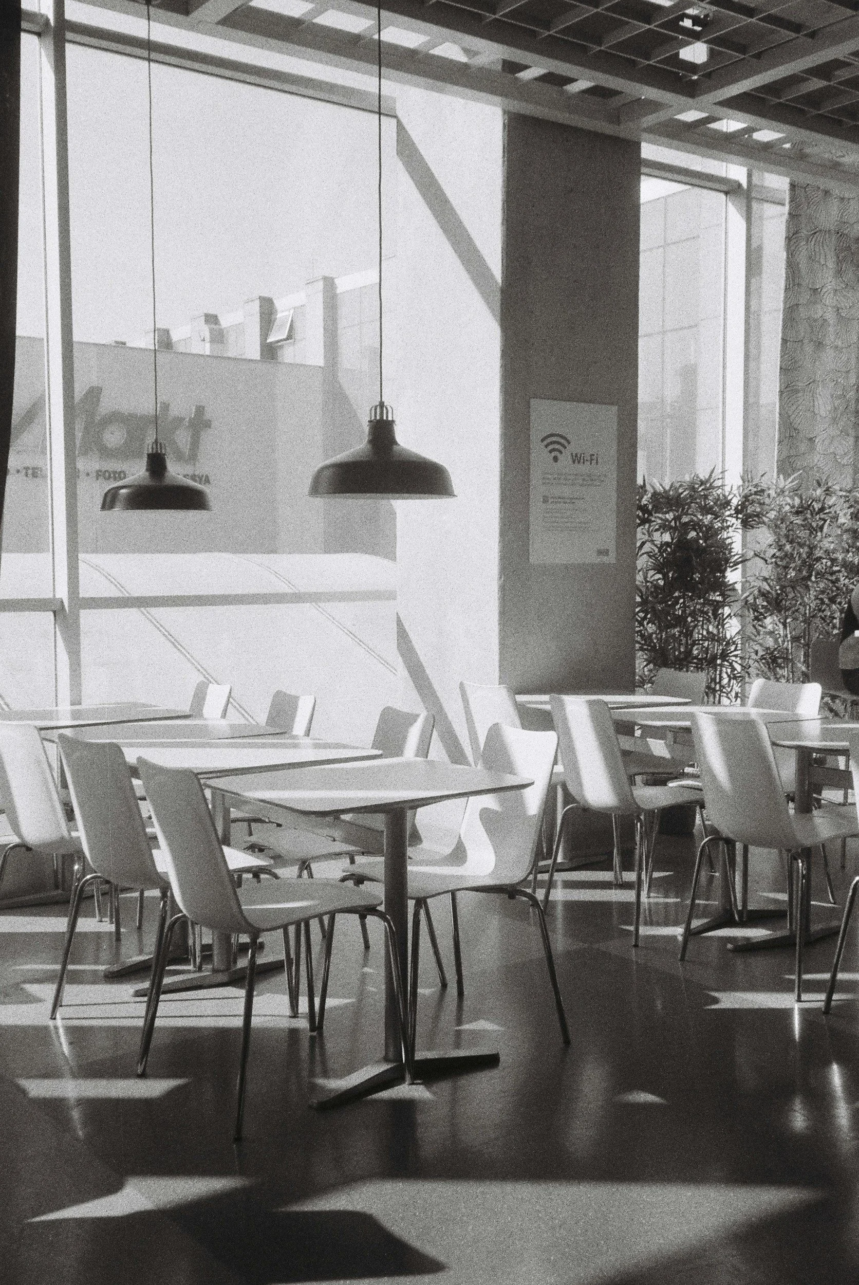 a grayscale image of a sunsoaked empty cafe, tables and white chairs near a window with pendant lights over the table