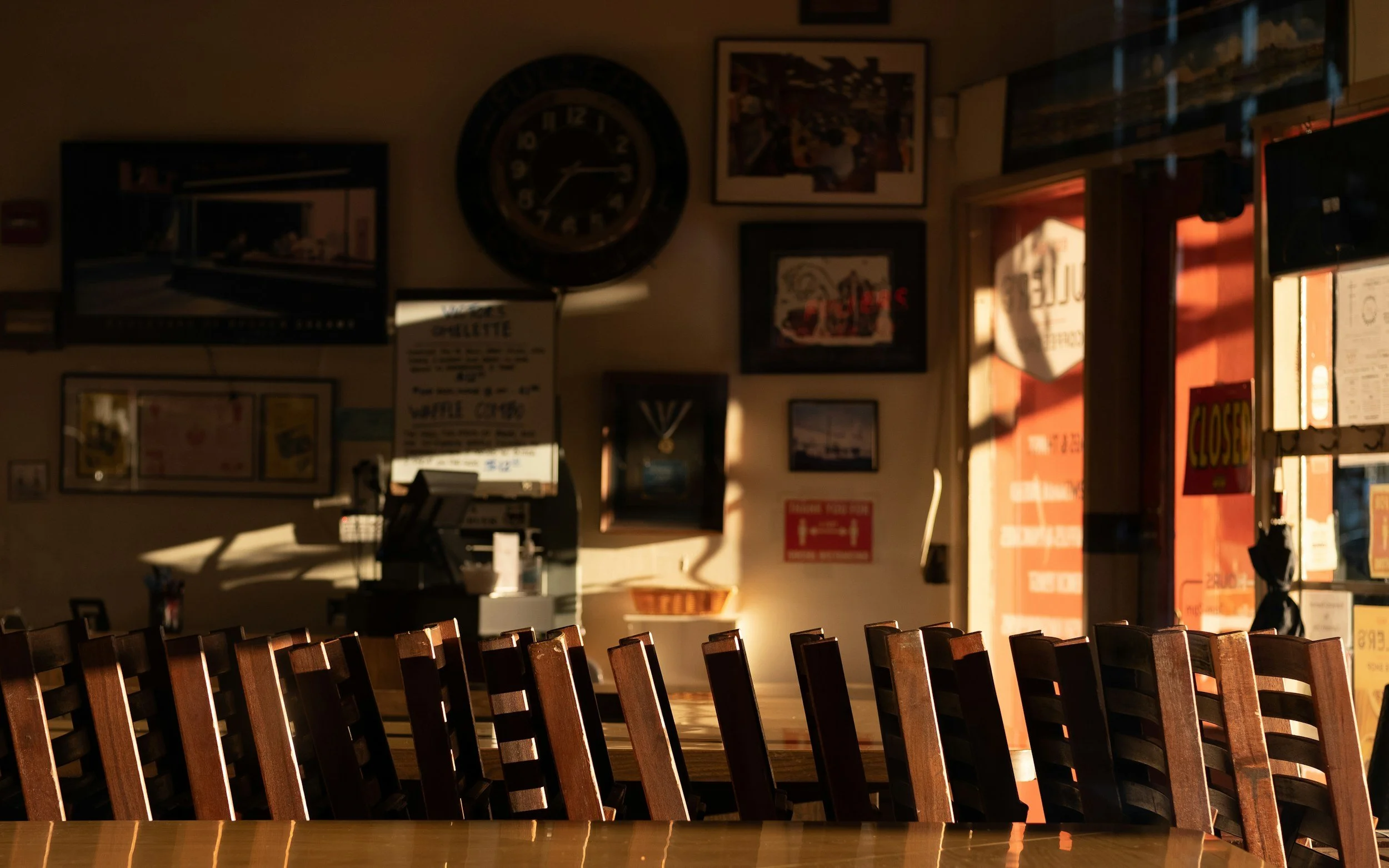 Inside a cozy restaurant or cafe, sunlight streams through the window, illuminating a row of wooden chairs and various framed pictures, posters, and a large clock on the wall.