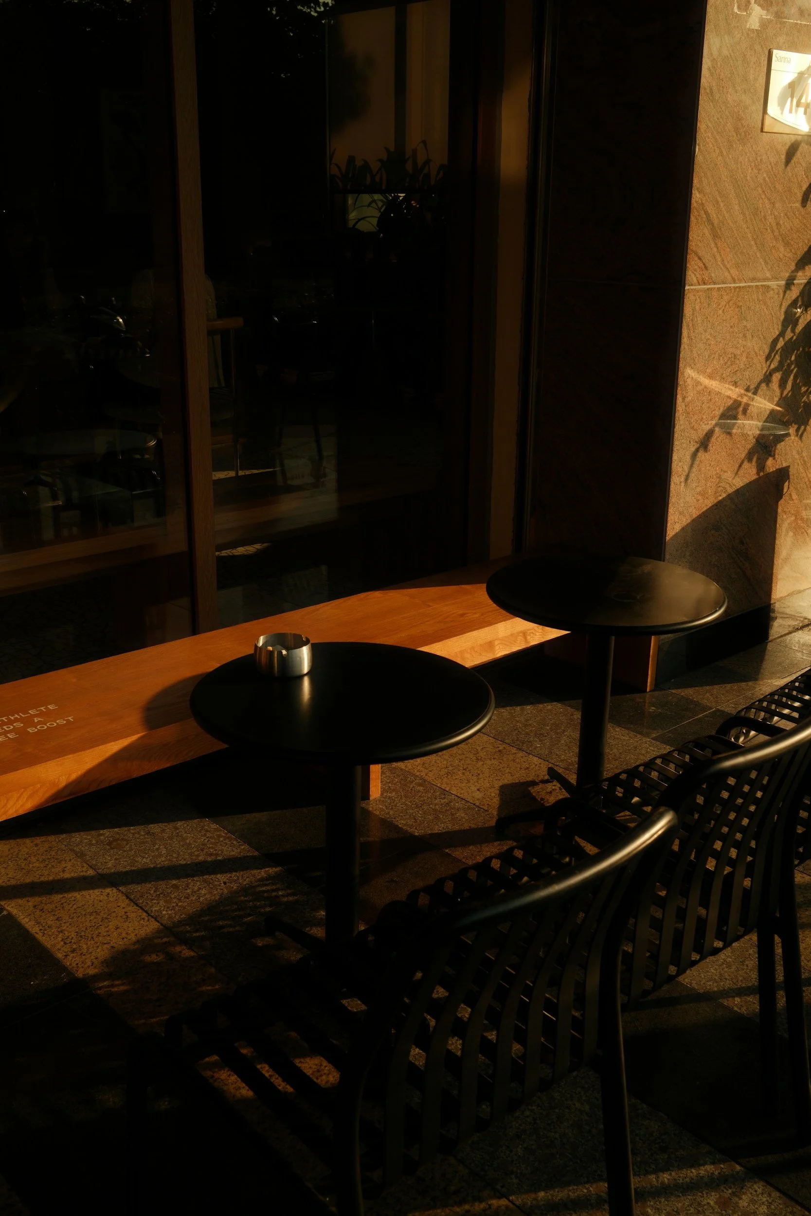 Outdoor scene at night with a wooden table, black chairs, and shadows cast by nearby plants on the wall.