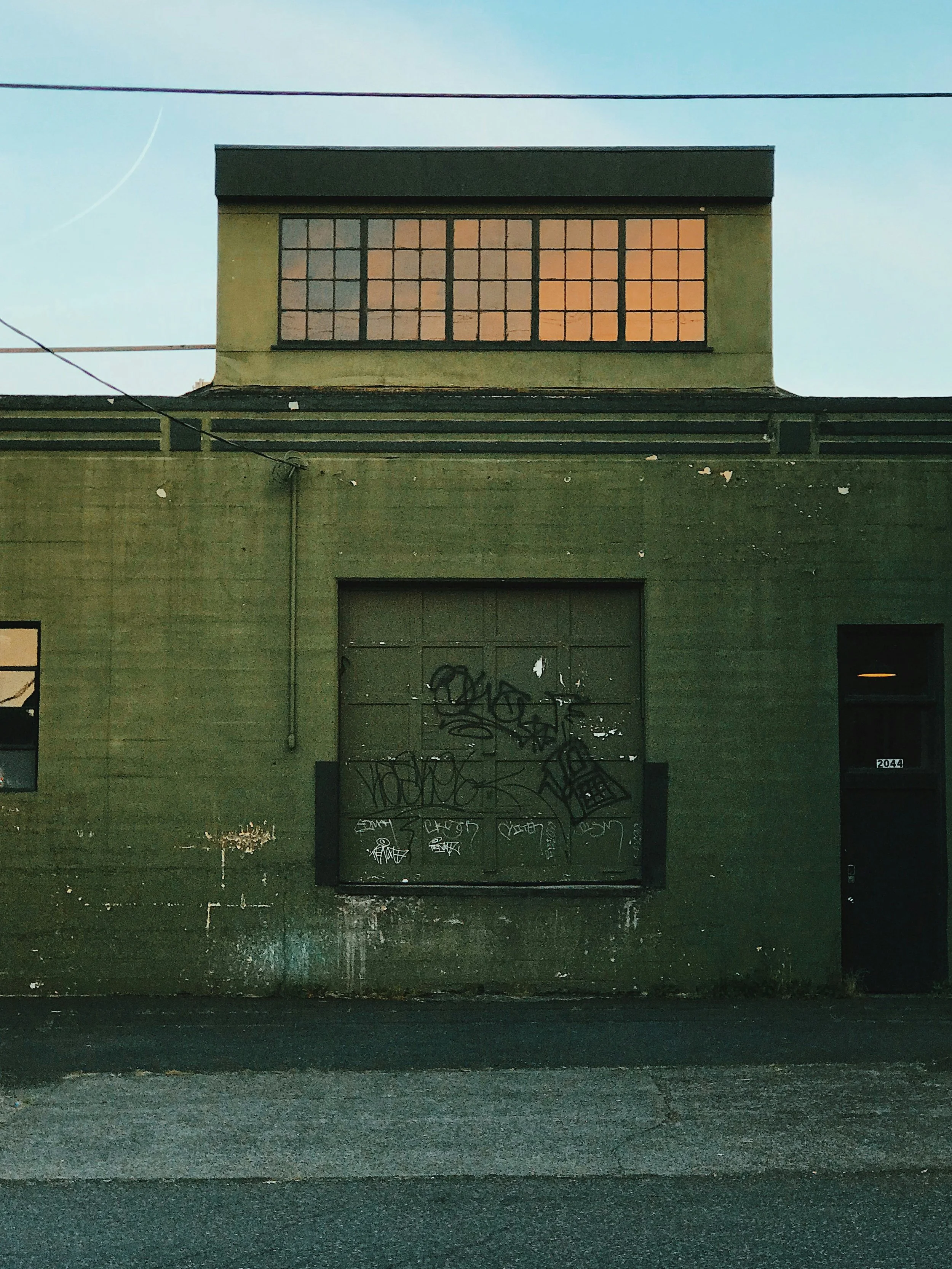The side of a green industrial building with patina and graffiti, with a set of windows on the second level reflecting a warm sunset