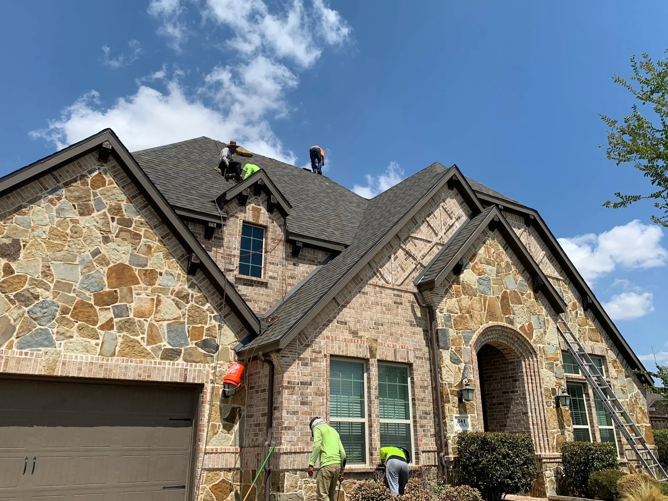 Three workers on the roof of a multi-story brick house, engaged in roofing work, under a partly cloudy blue sky, with lush green tree on the right side.