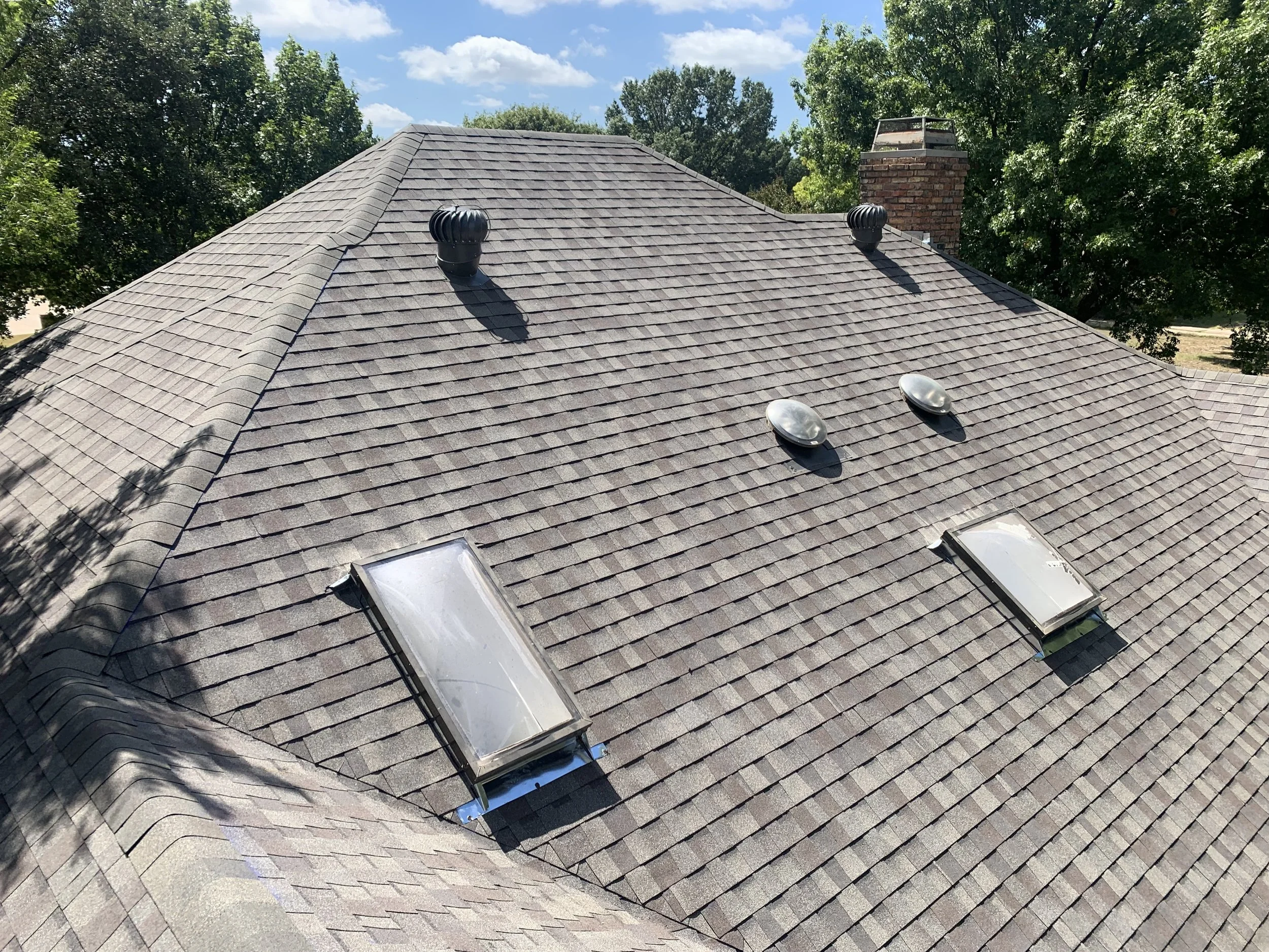 A roof with asphalt shingles, two skylights, three vents, and a brick chimney, surrounded by green trees and a blue sky with clouds.