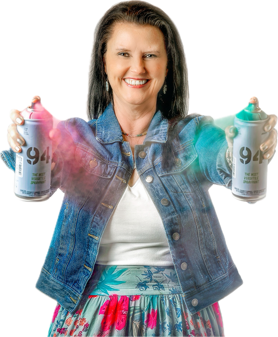 A woman with dark hair, smiling, wearing a denim jacket, a white top, and a colorful floral skirt, holding spray paint cans in both hands, with colorful spray paint in the air.