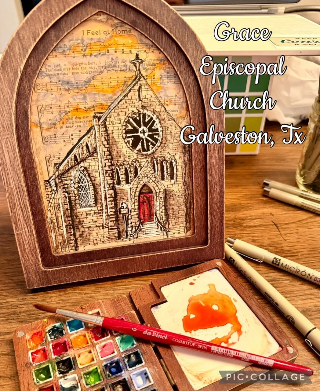 A framed artwork of a church with musical notes, surrounded by art supplies including watercolors and pens, on a wooden table.