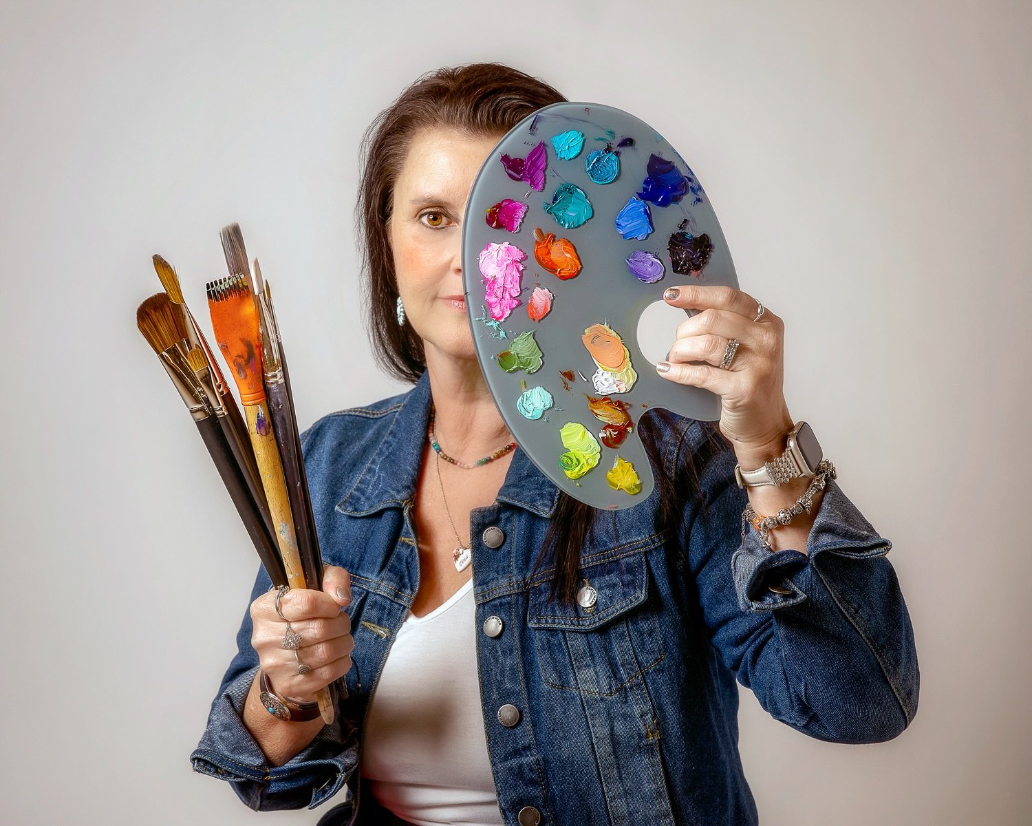 A woman holds a painter's palette with various colors of paint and artist brushes, partially obscuring her face, against a light background.